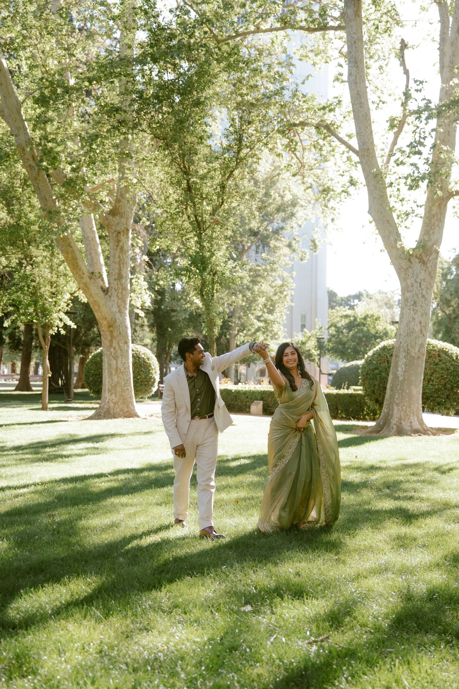 Couple dancing outdoors in a lush green park, woman in olive saree and man in white suit, trees in background.