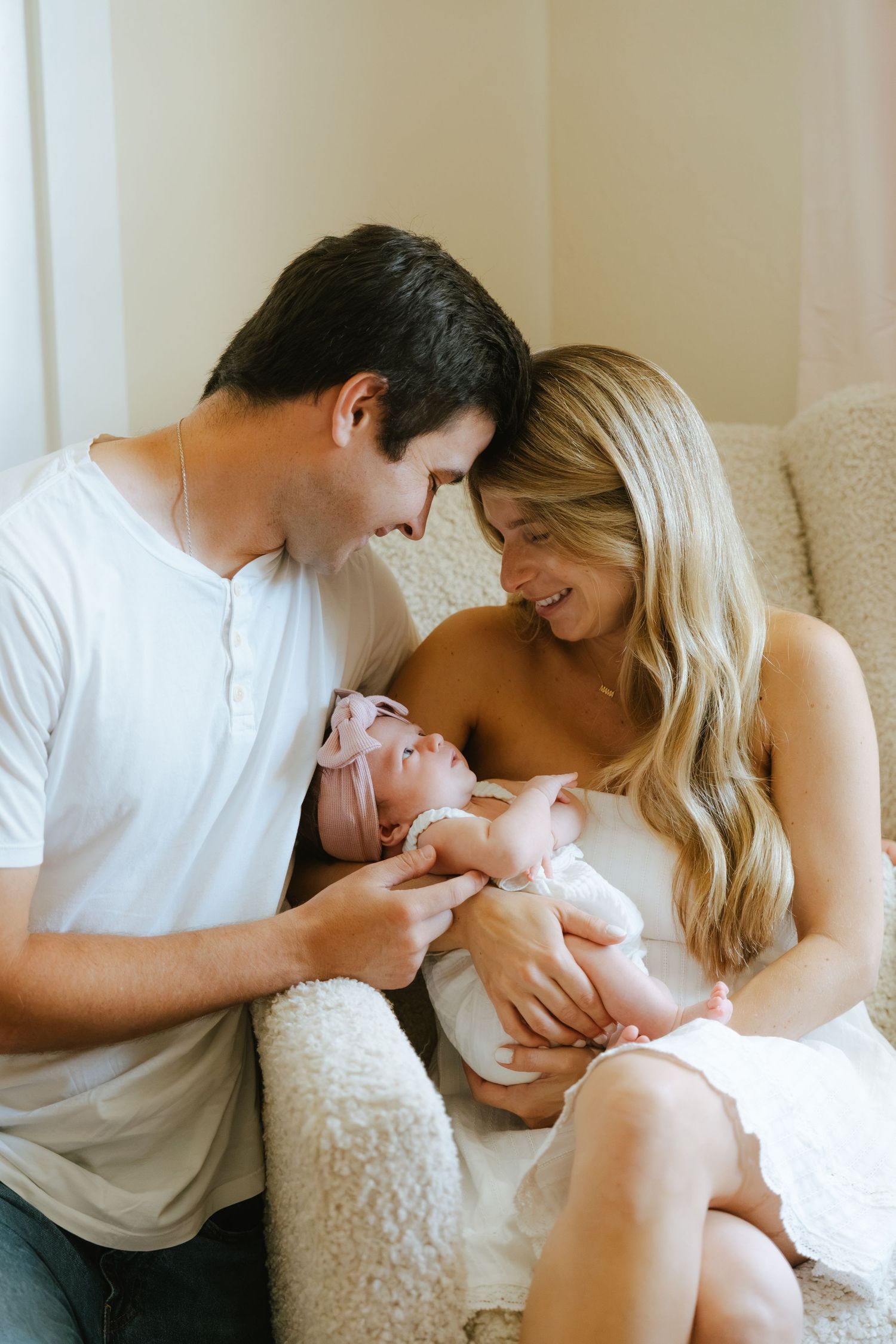A couple smiles lovingly at their newborn baby wrapped in white with a pink headband while sitting on a cozy chair.