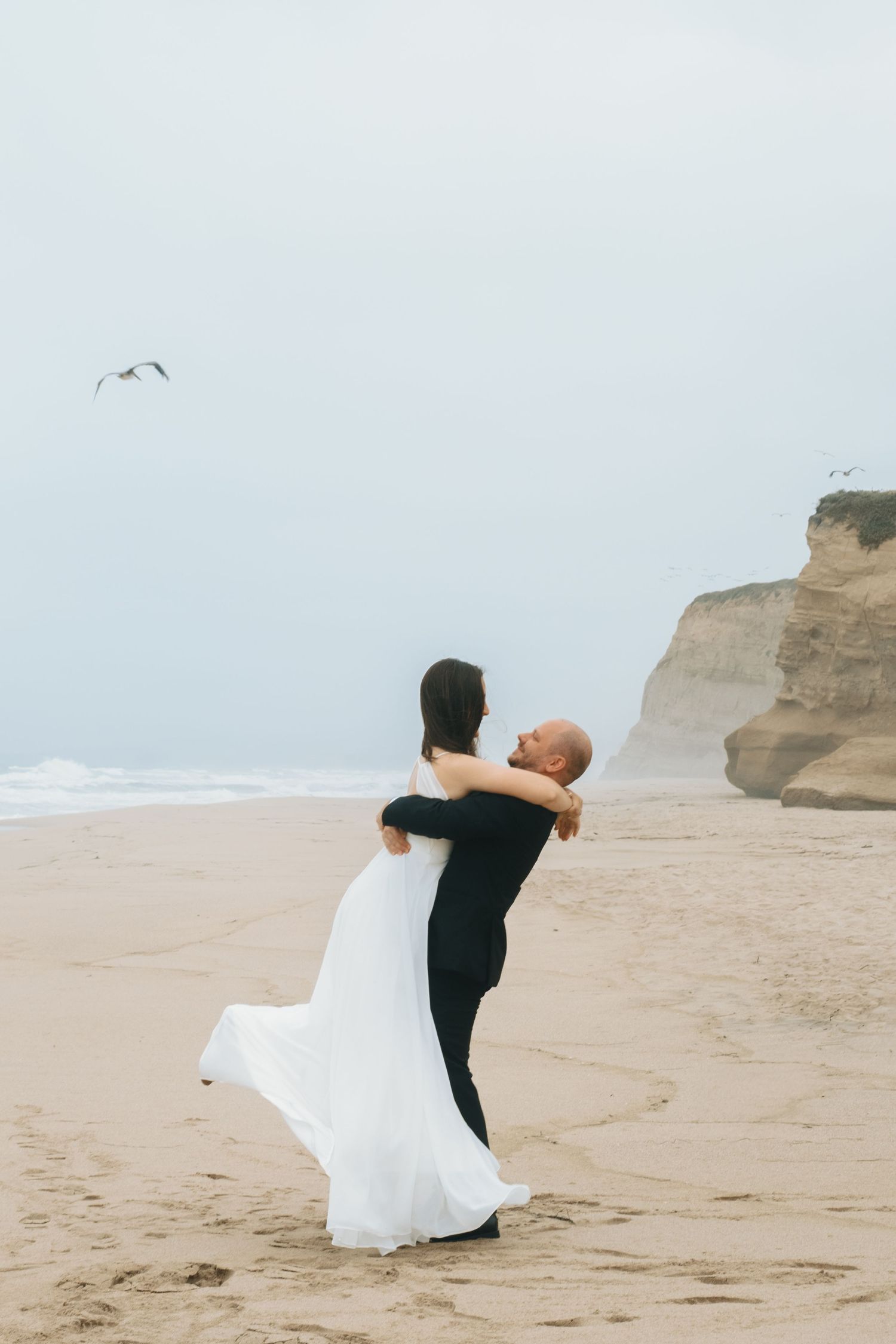 A groom lifts his bride in a white gown on a misty beach, with ocean waves and rocky cliffs in the background.