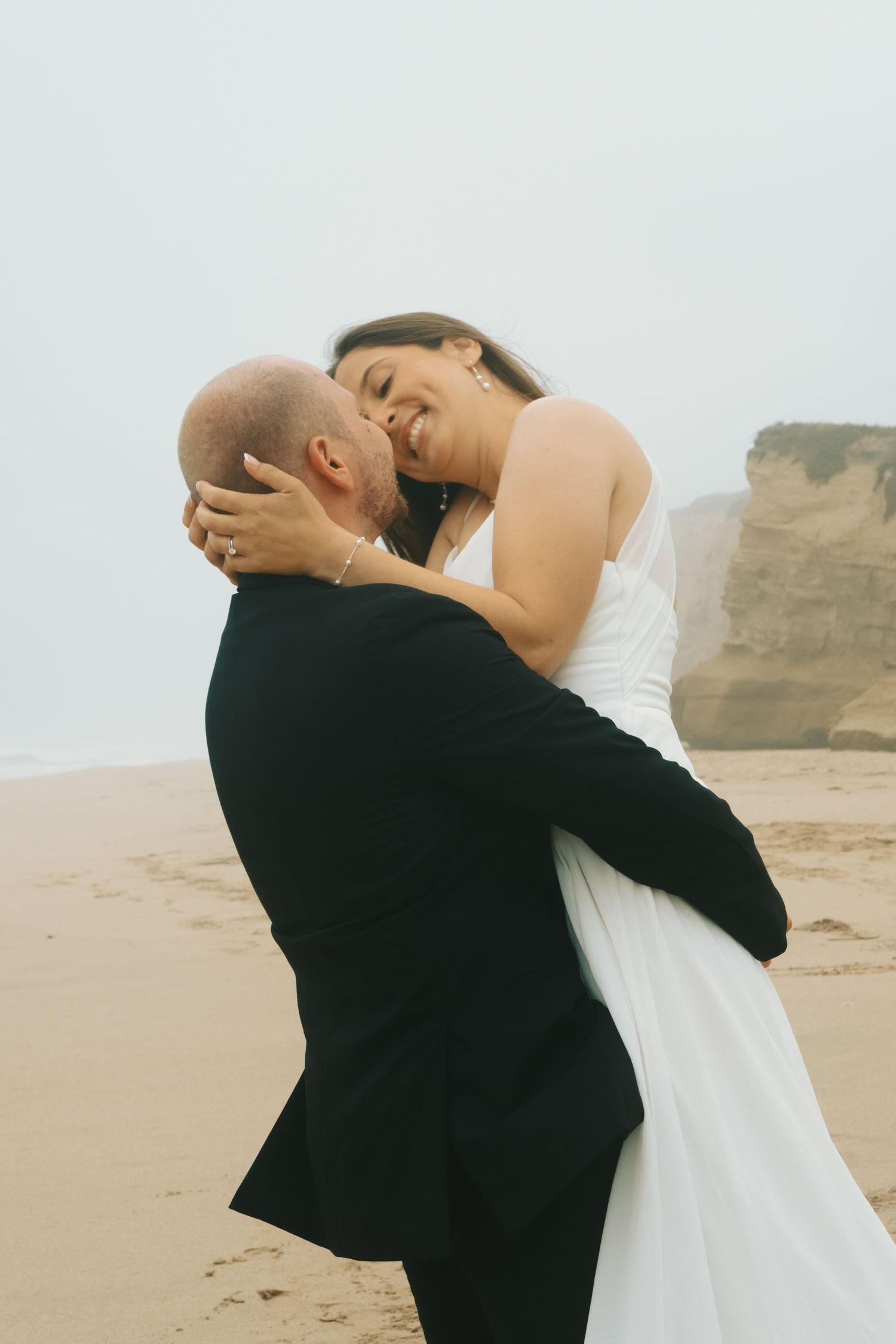 A joyful bride in a white dress embraces a bald groom in a black suit on a sandy beach with misty cliffs.