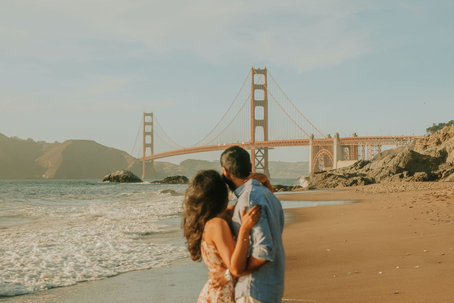 A romantic beach photoshoot with the iconic Golden Gate Bridge visible in the background during sunset.