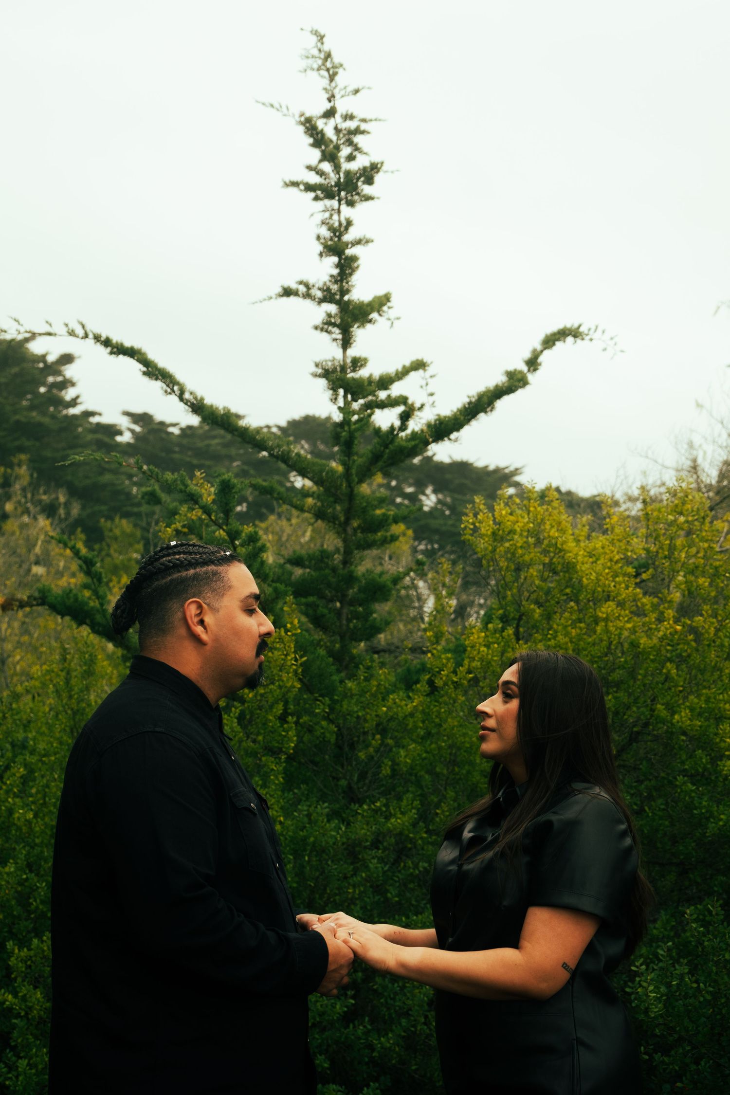 Couple having animated conversation outdoors with evergreen trees and yellow flowers in background.