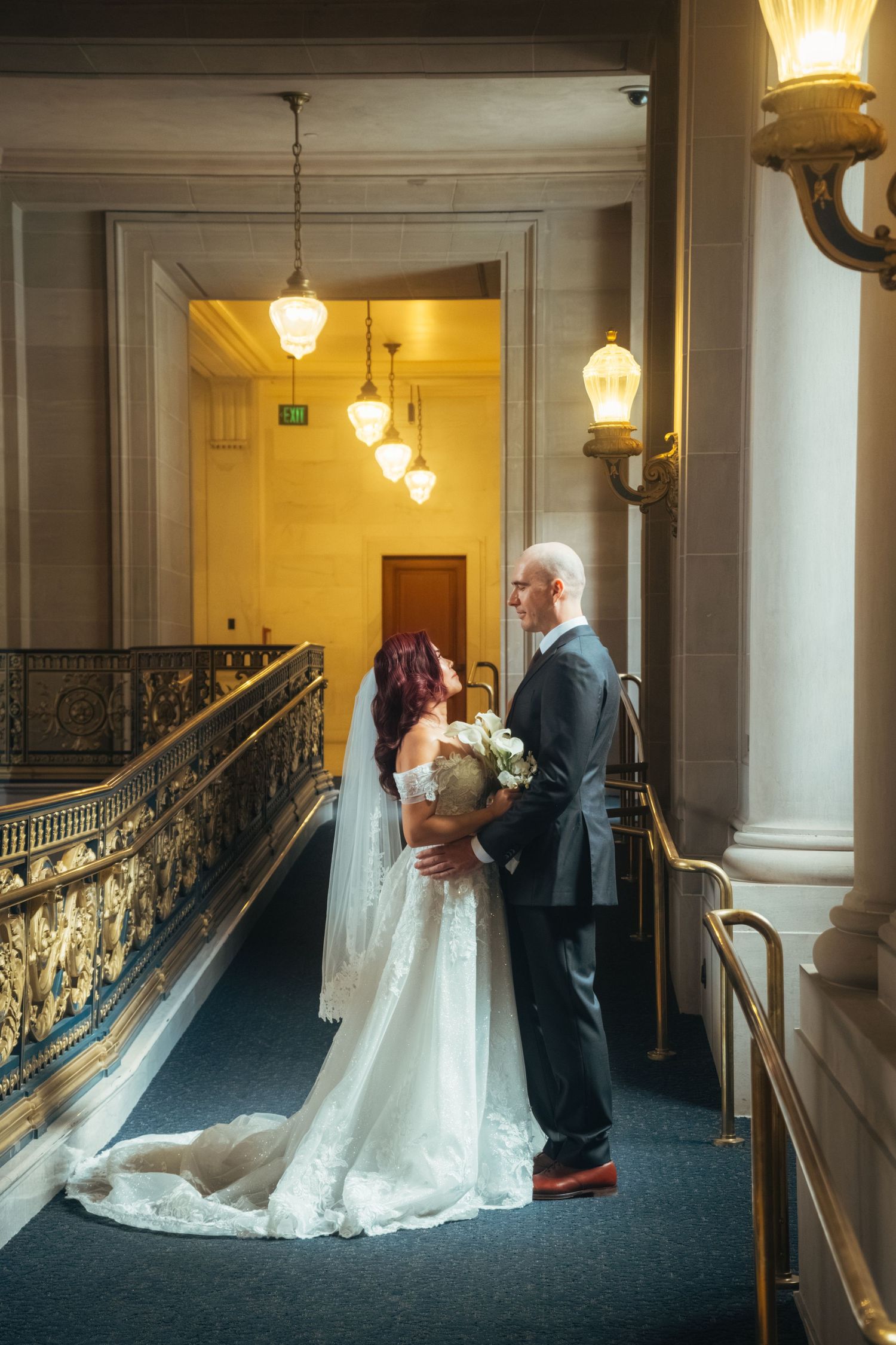 Wedding couple embracing in elegant hallway with vintage lighting and ornate railings.