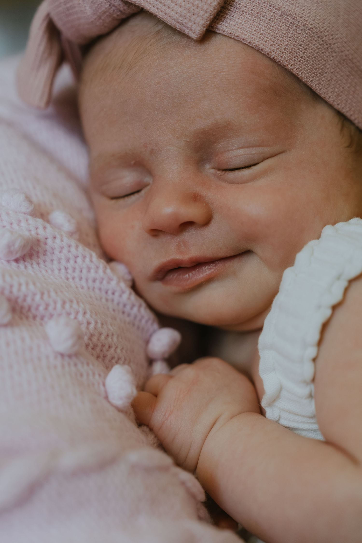 Sleeping newborn baby girl in pink knit hat and white outfit, peacefully resting on a soft pink blanket.