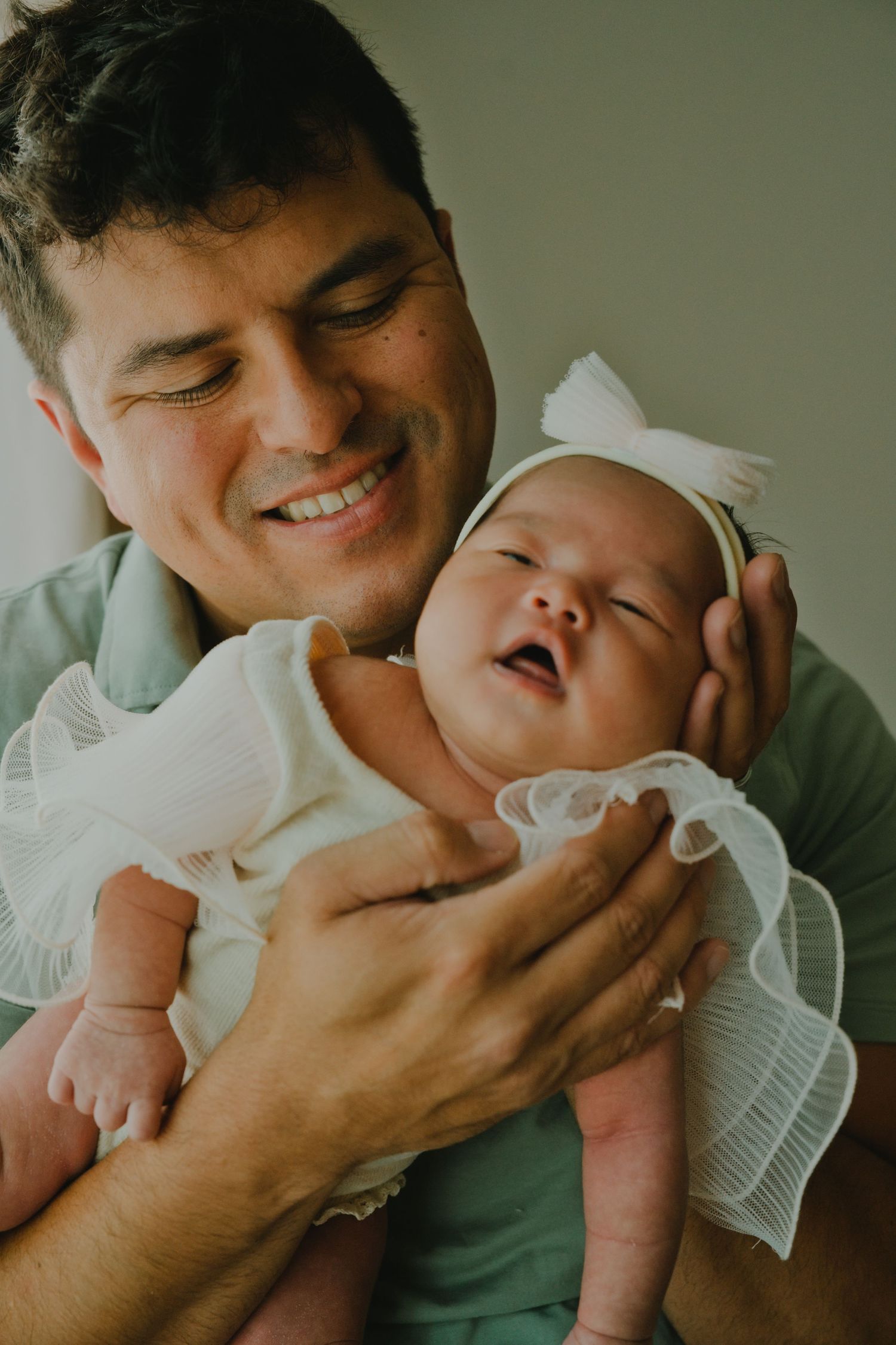 Smiling father holding his newborn baby girl dressed in white with a bow headband.
