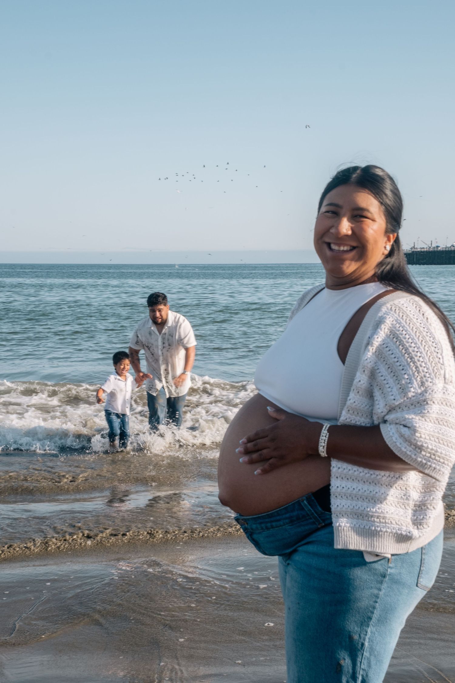 Smiling pregnant woman on beach in white top and jeans, with man and child playing in ocean waves behind her.