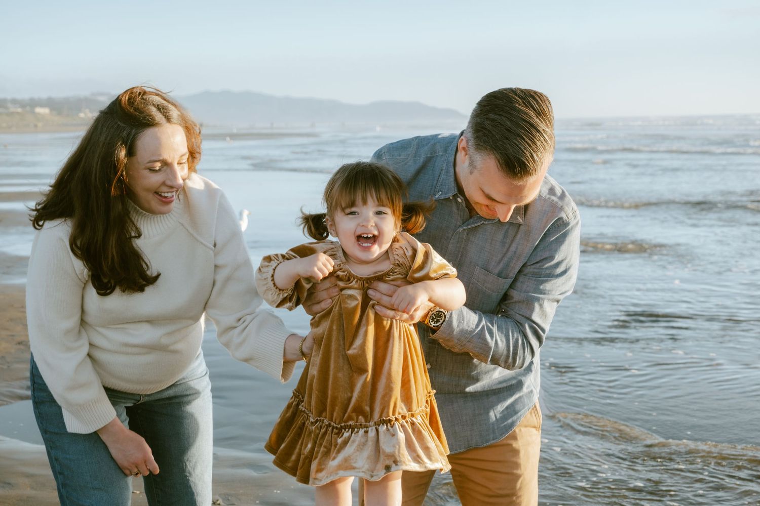 Family of three enjoying a sunny beach day, parents helping their laughing toddler girl walk along the shoreline.