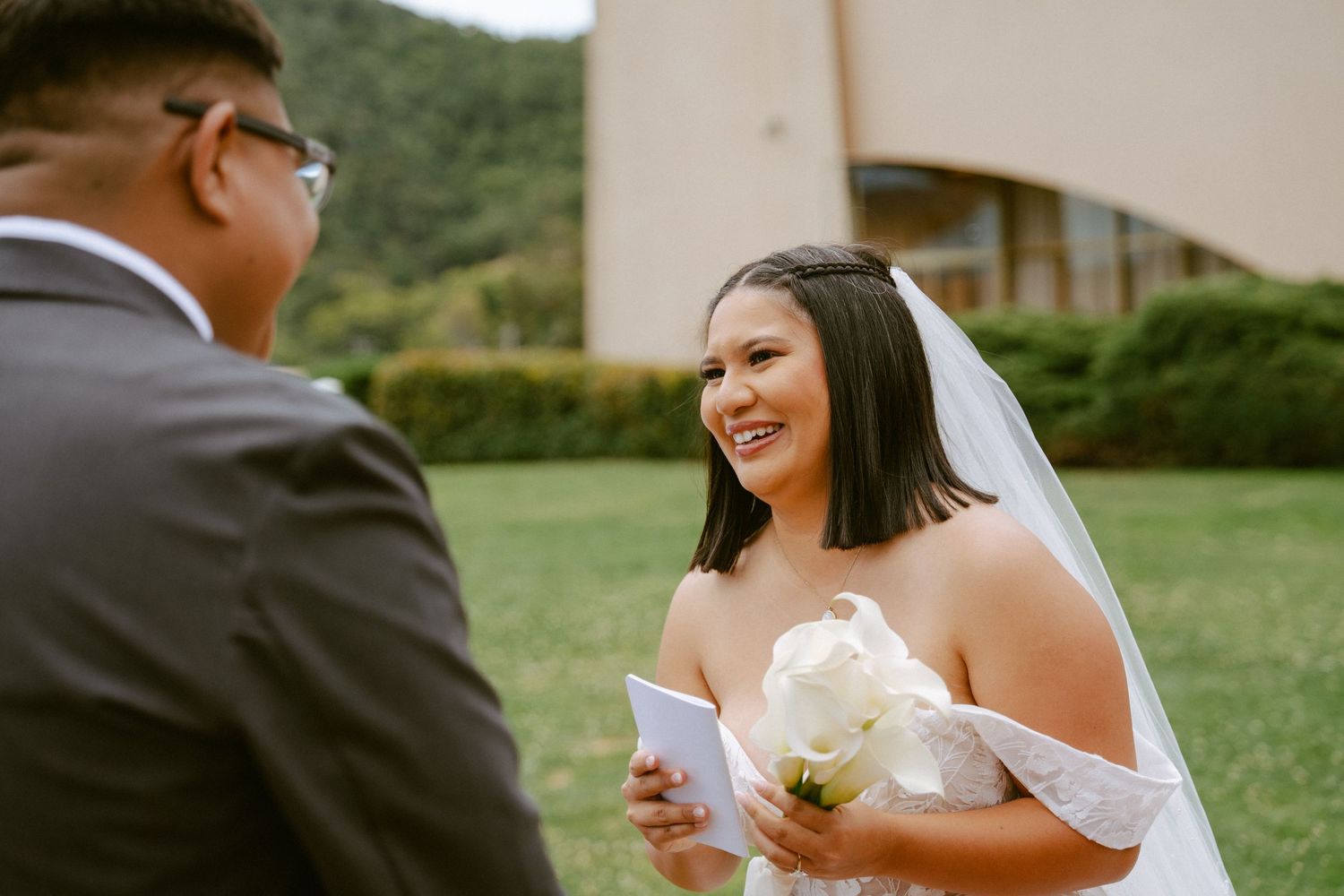 Smiling bride in white lace gown and veil holds bouquet and vows card during outdoor wedding ceremony.