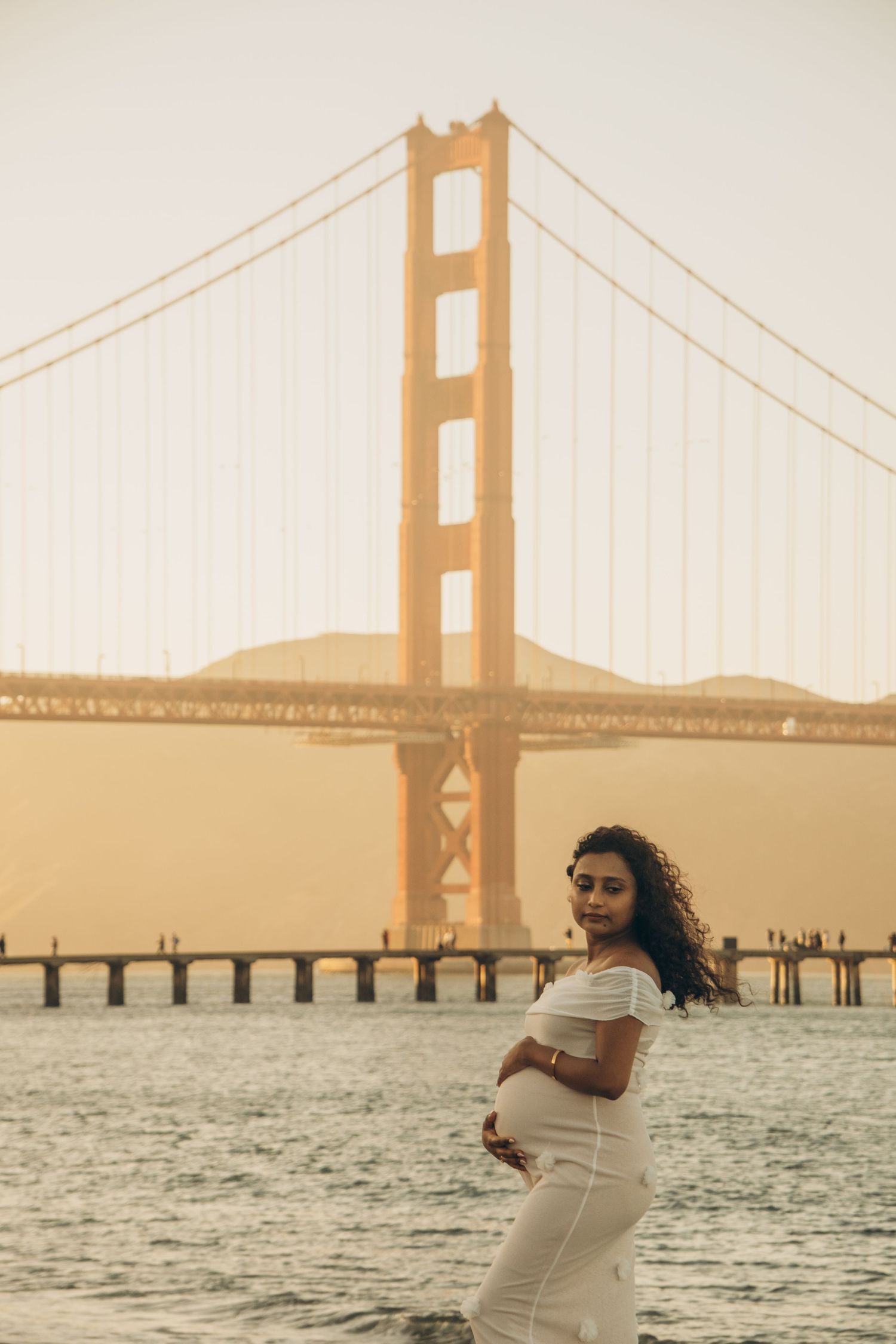 Woman in white dress poses near Golden Gate Bridge at sunset.