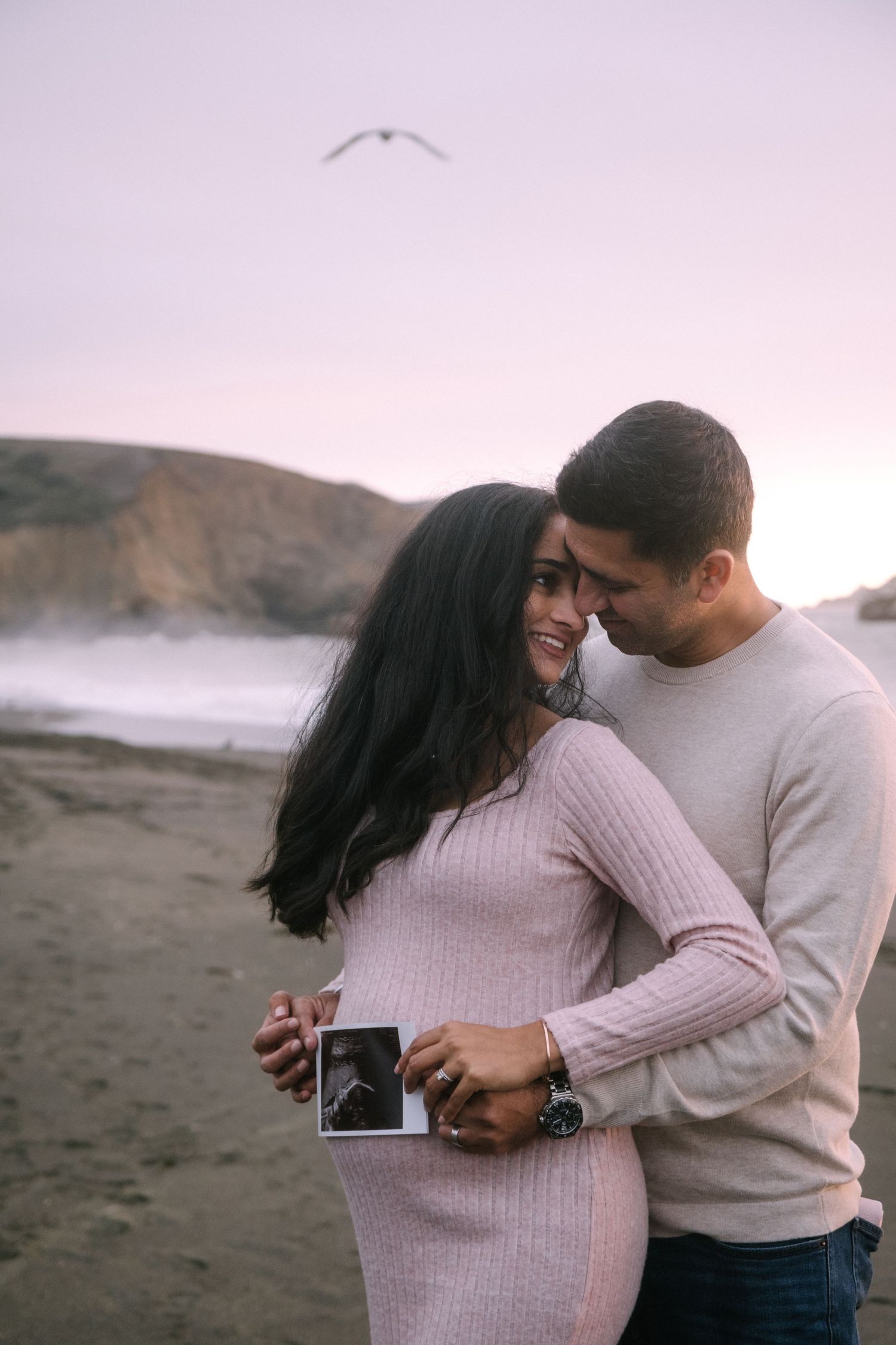 Couple examining something together on a beach with soft pink sky overhead.
