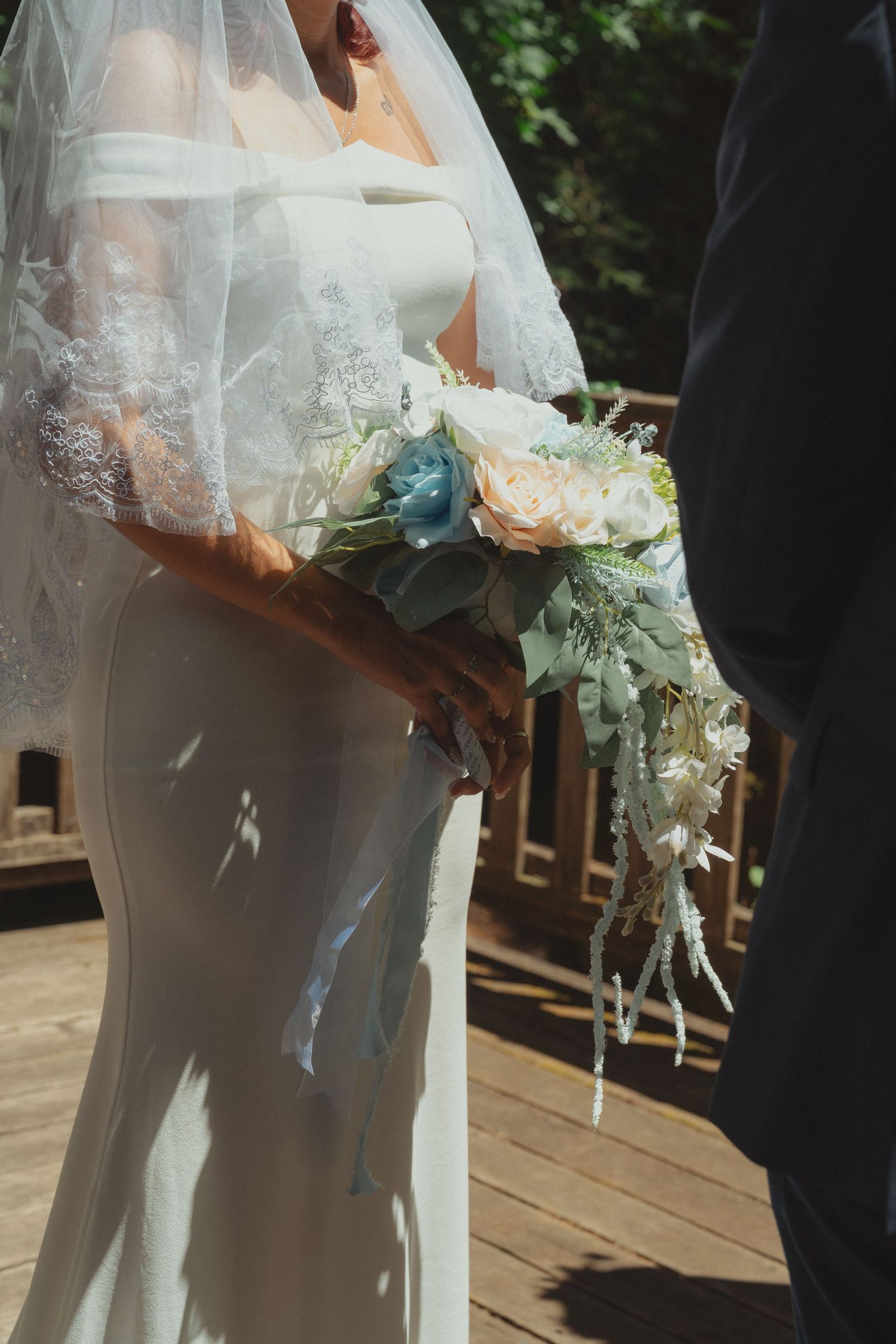 Bride in white gown and veil holding a bouquet of peach and blue flowers on a wooden deck.