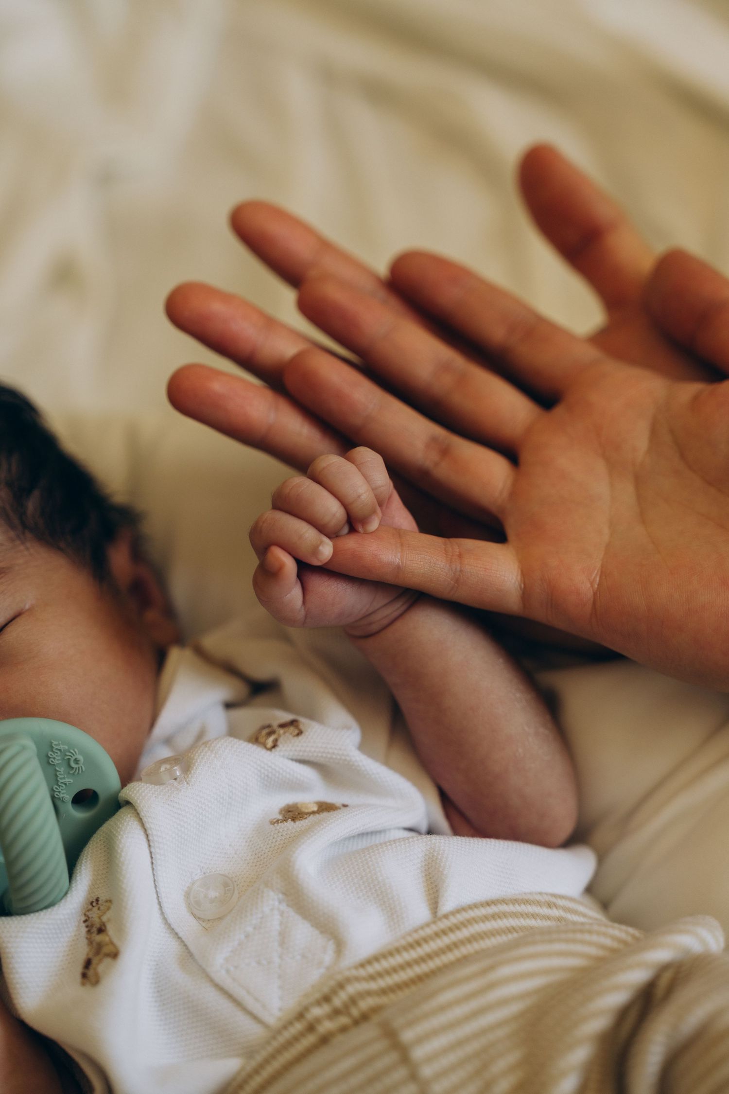 Newborn baby sleeping and gripping an adult's finger, wearing a white onesie with small prints on a beige background.