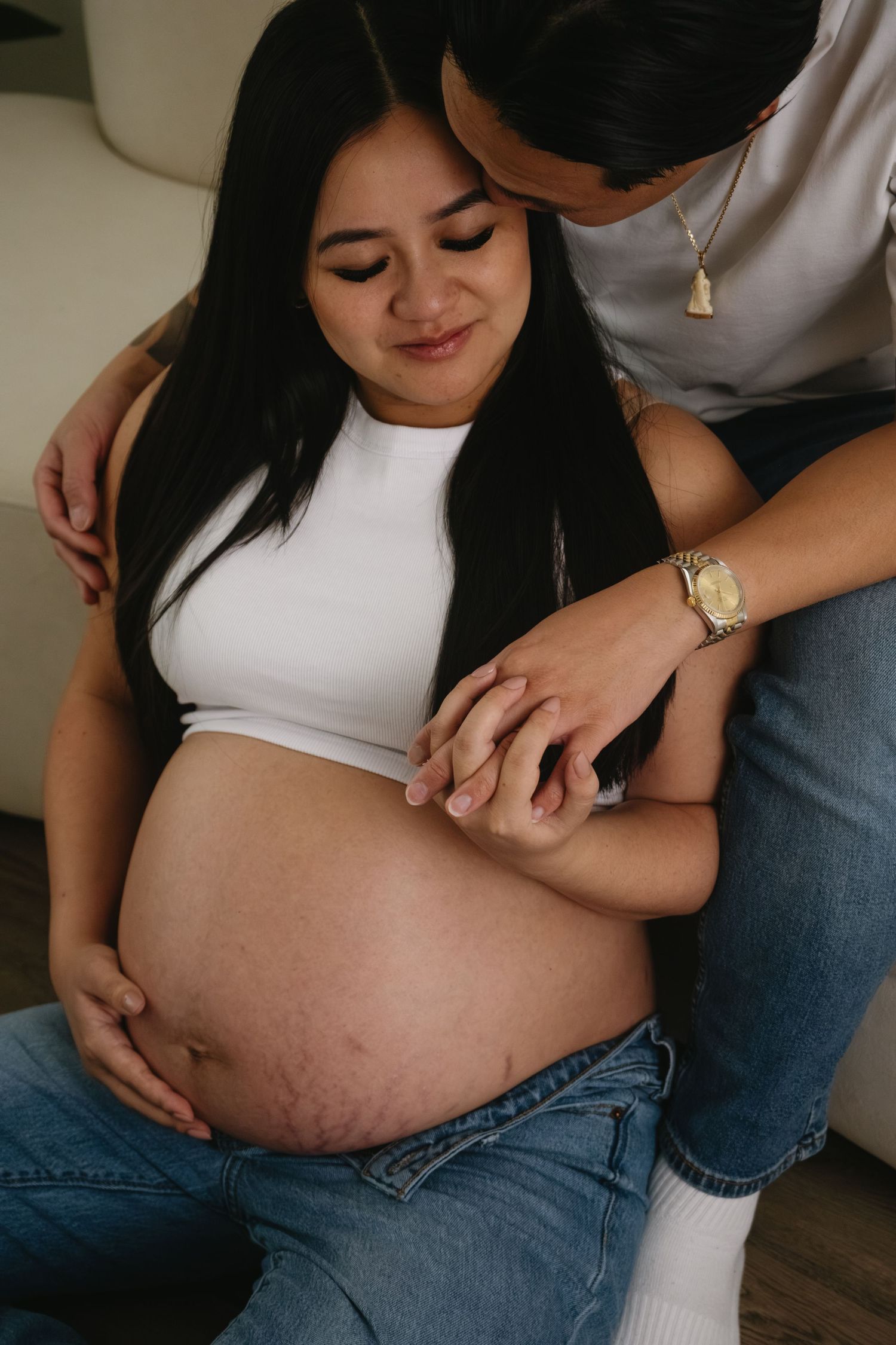 Intimate maternity photo showing a pregnant woman in white crop top being embraced while sitting on floor.