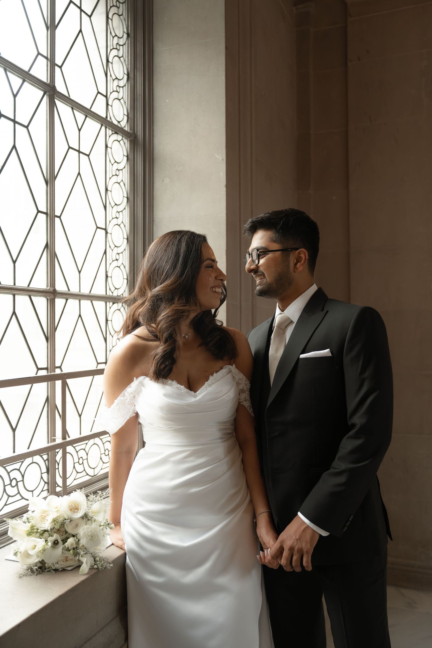 Bride and groom holding hands by ornate window, she in white off-shoulder gown, he in black suit, sharing a loving gaze.