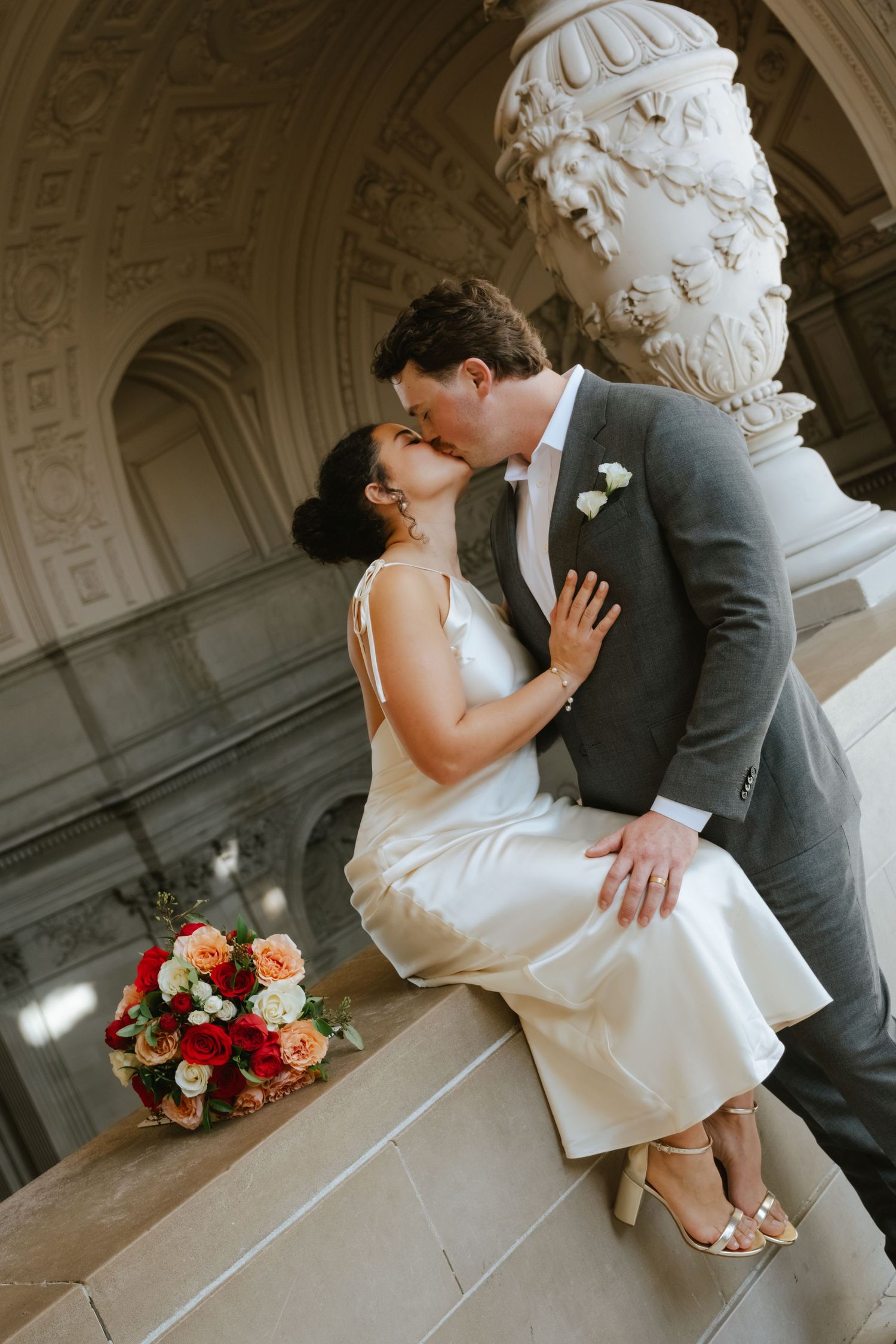 Bride and groom share a romantic kiss on marble steps, with a red and peach rose bouquet, at an elegant venue.