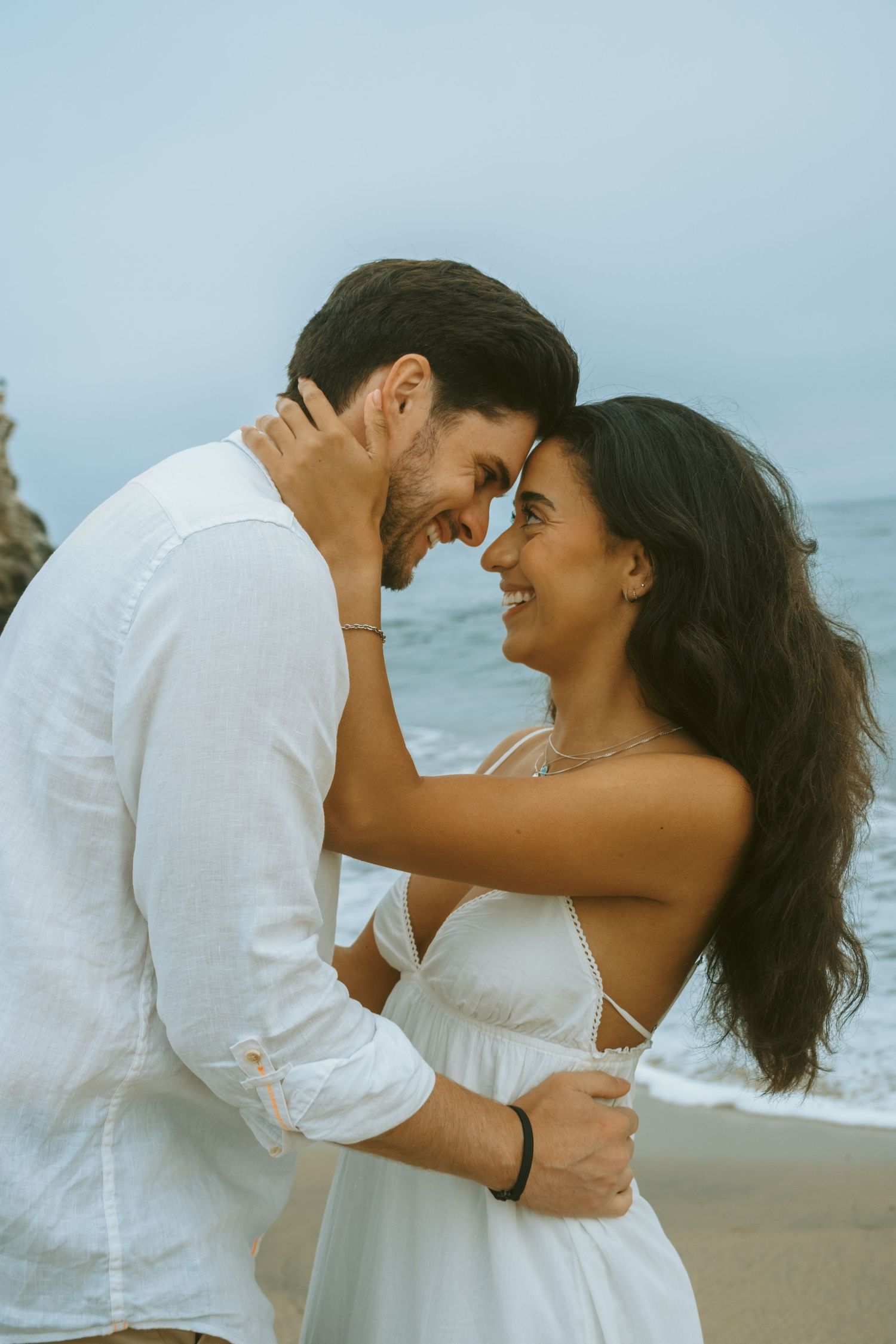 A tender moment between a couple in white attire sharing an embrace on the beach.