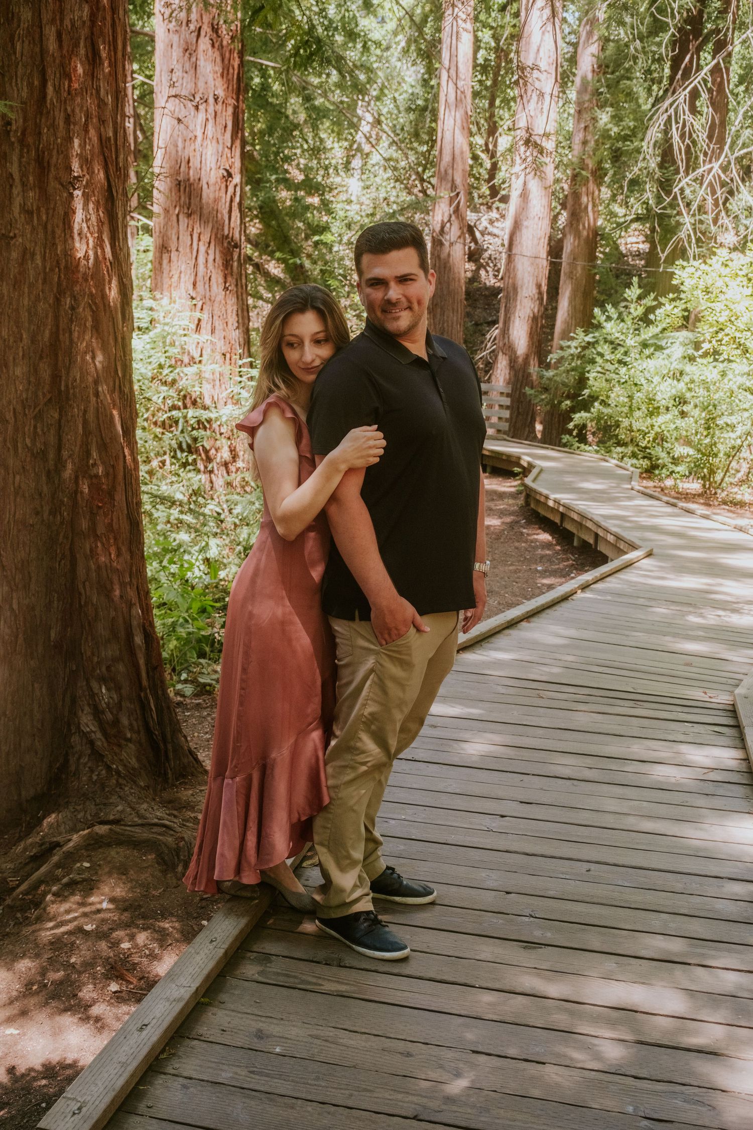 A couple shares playful moments together on a wooden walkway through a redwood forest.
