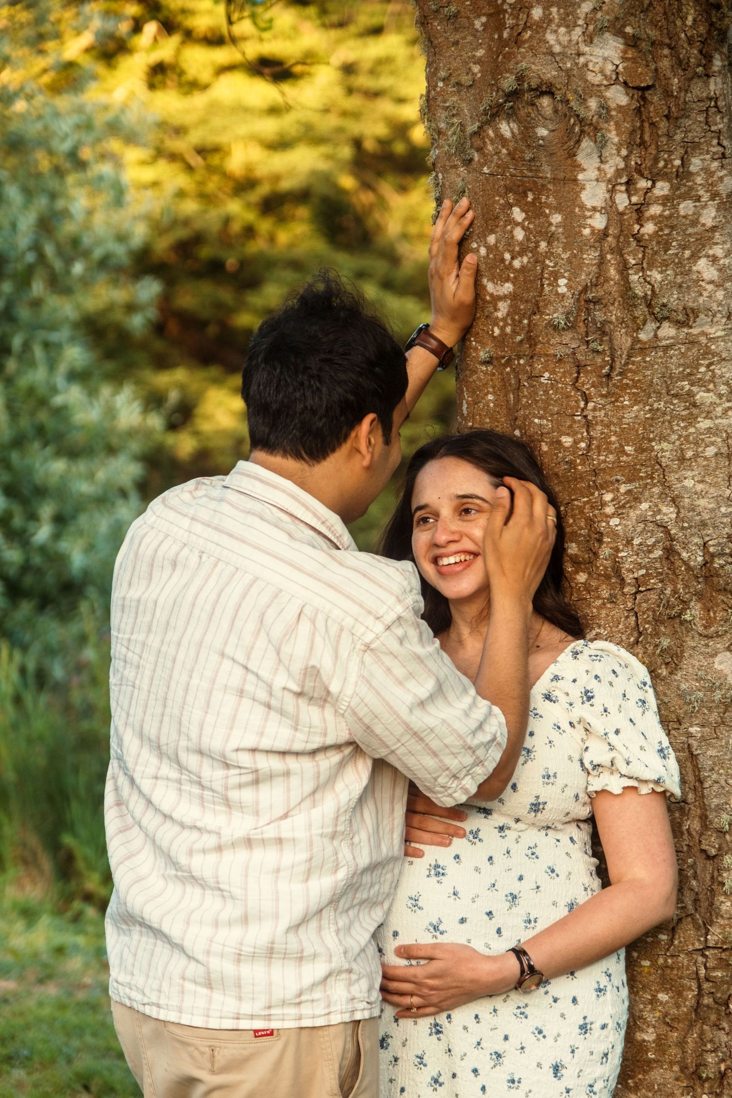 Romantic couple sharing intimate moments by a tree trunk in a sunlit forest setting.