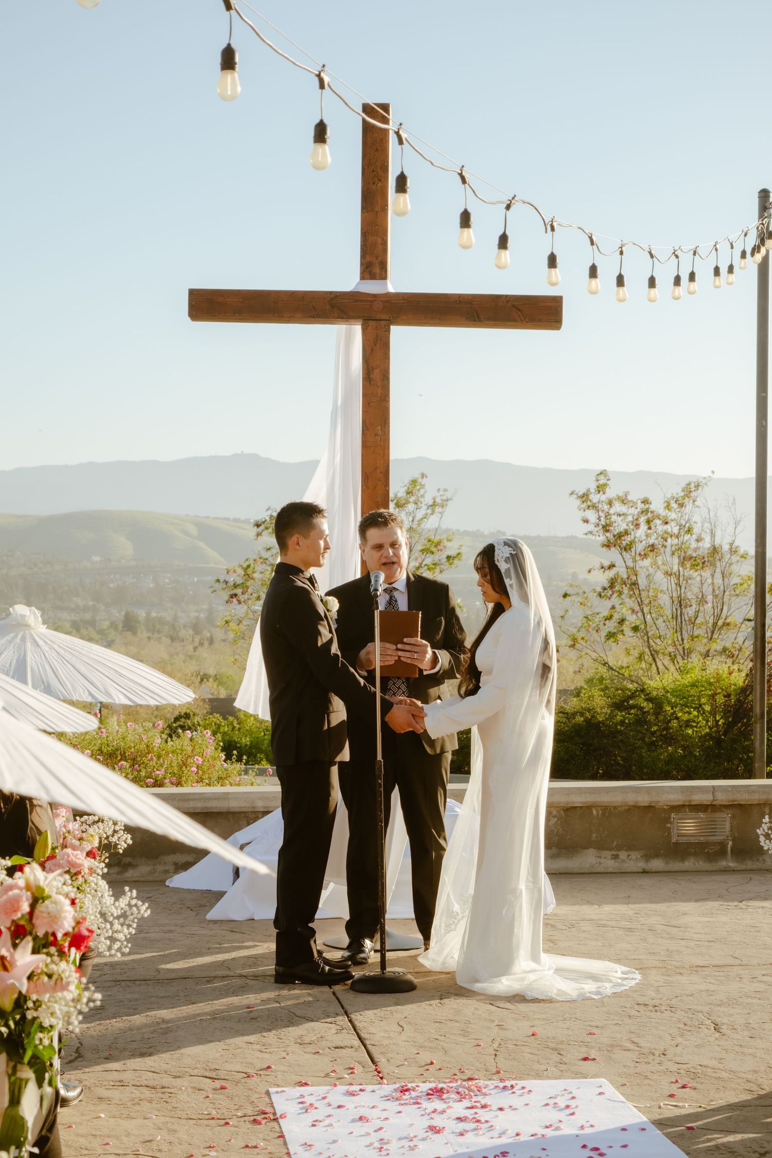 Couple exchanges vows at outdoor wedding ceremony before a wooden cross with scenic mountain views.