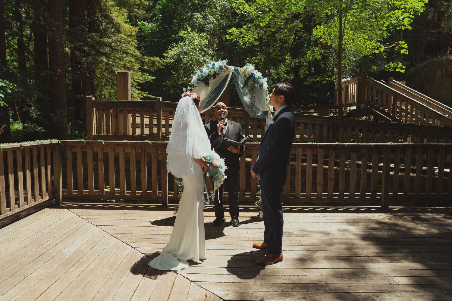 Bride and groom exchange vows on a wooden deck surrounded by lush redwood forest during an outdoor wedding ceremony.