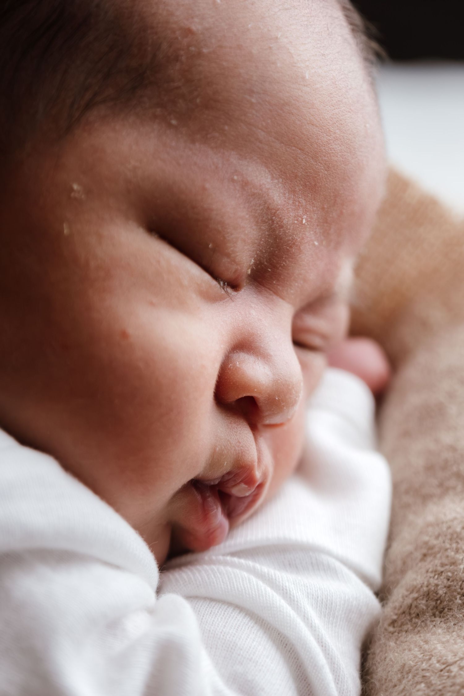 Close-up of a sleeping baby wearing white clothing against a soft brown blanket.