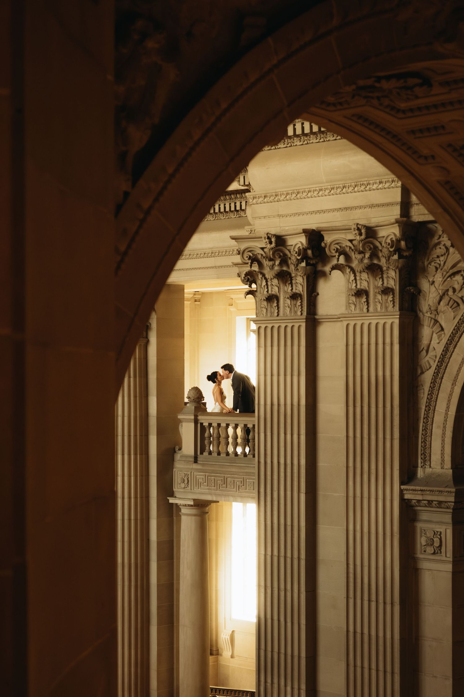 Ornate classical architecture with Corinthian columns and arches frames a silhouetted couple on a balcony in warm lighting.
