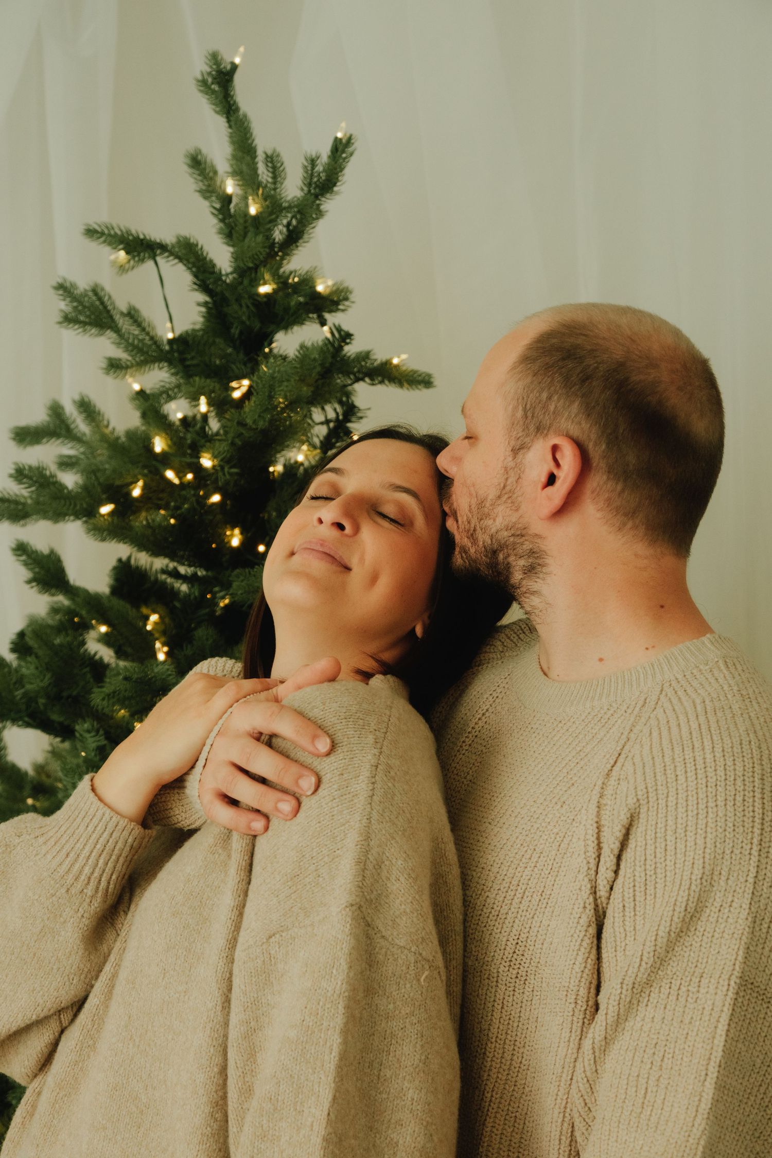 Tender embrace captured in front of lit Christmas tree with couple in matching beige sweaters.