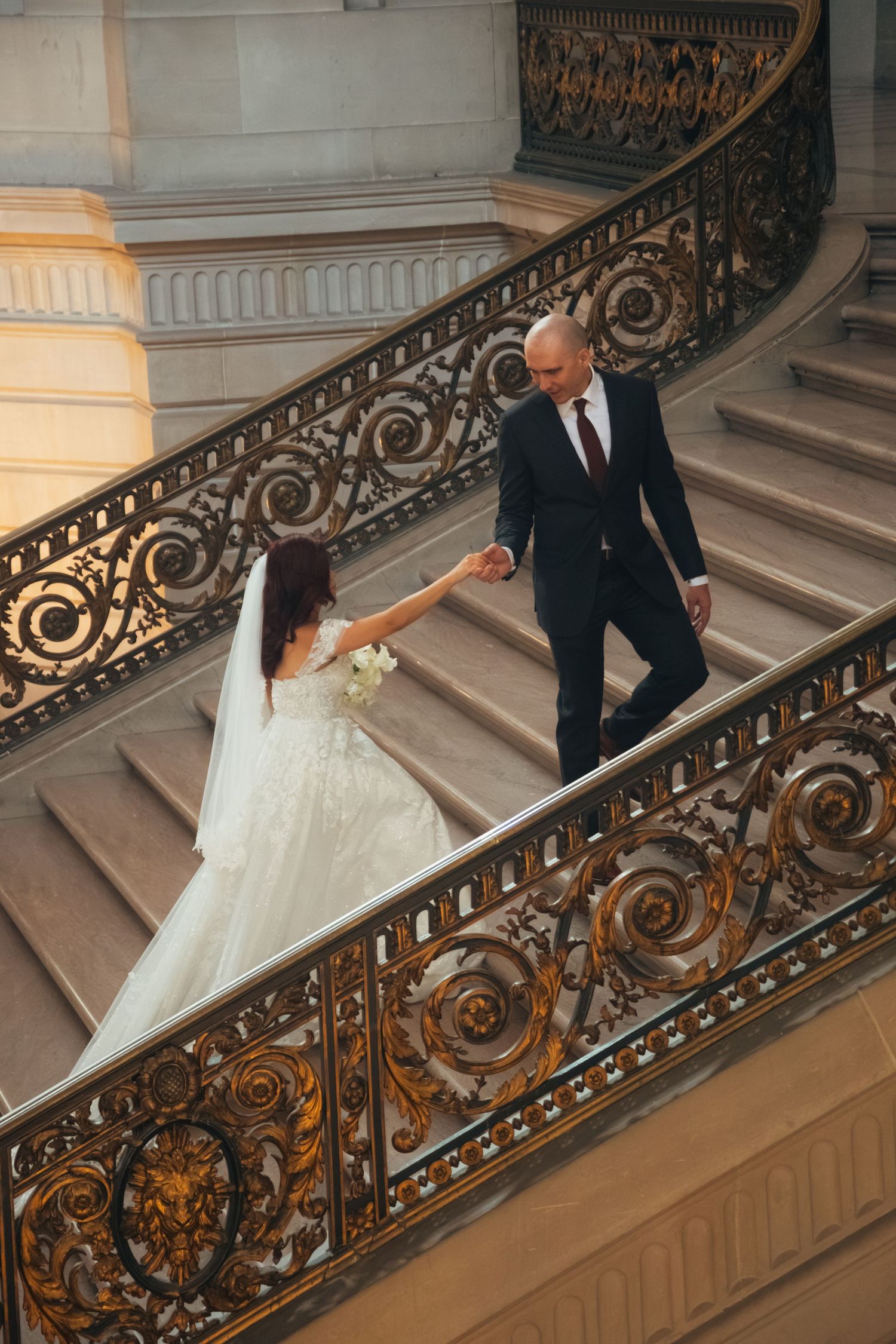 Couple descending ornate staircase with detailed wrought iron railing in historic building.