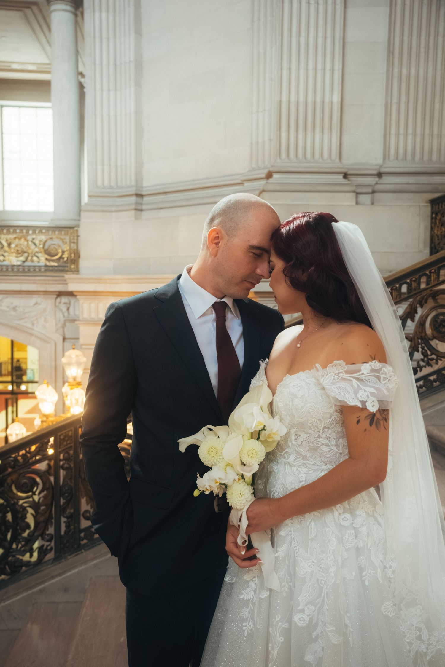 A romantic wedding couple shares an intimate moment on a grand marble staircase in an elegant venue.