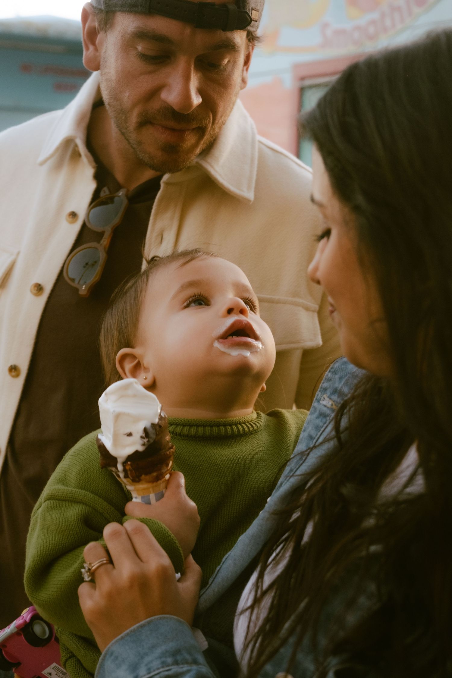 A baby in a green sweater holds an ice cream cone while being held by two smiling adults outdoors.
