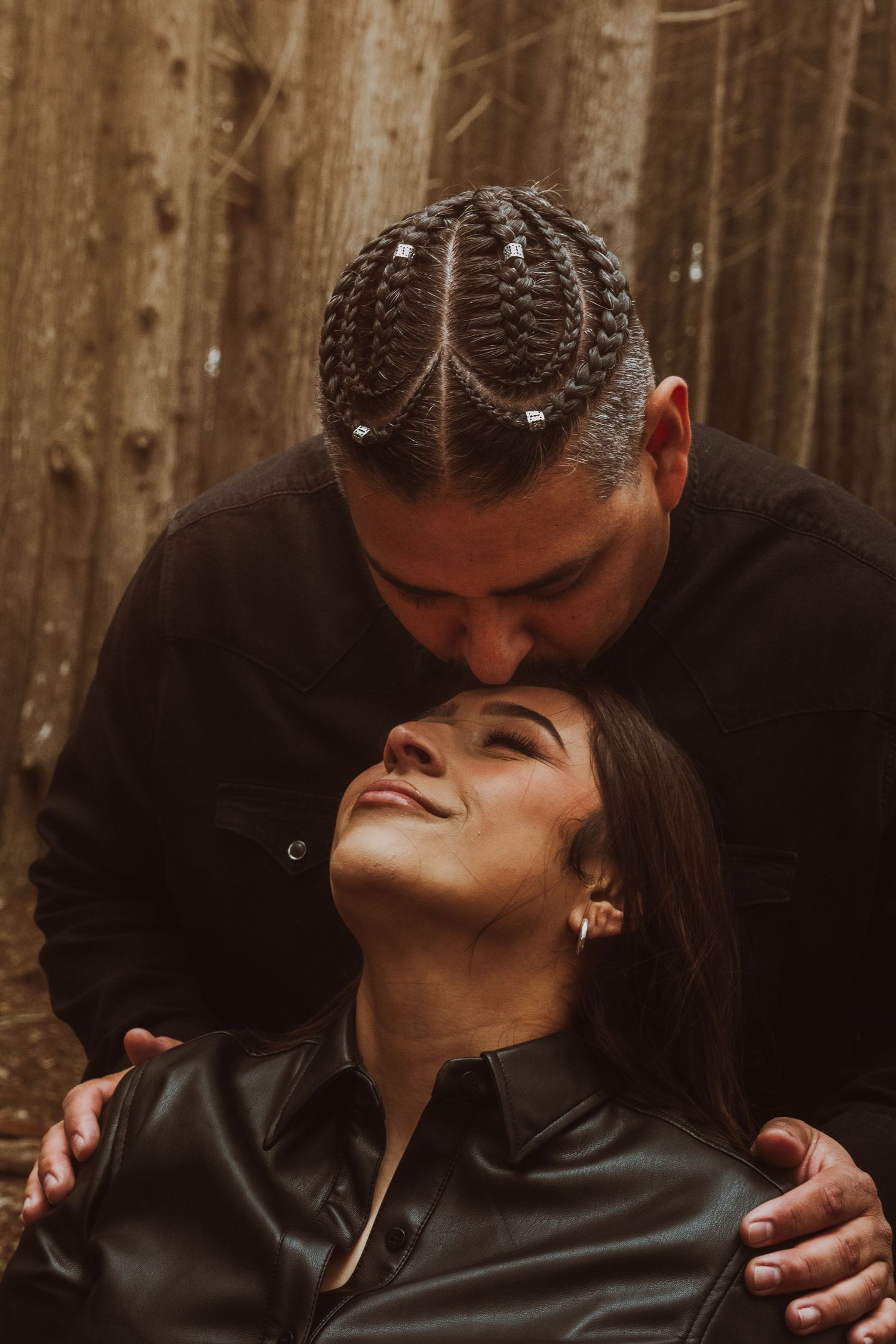 A man with cornrow braids kisses a smiling woman on the forehead in a romantic forest setting.