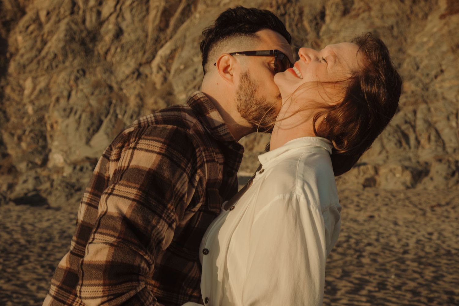 Romantic couple sharing a tender kiss on a sandy beach at golden hour with rocky cliffs in the background.