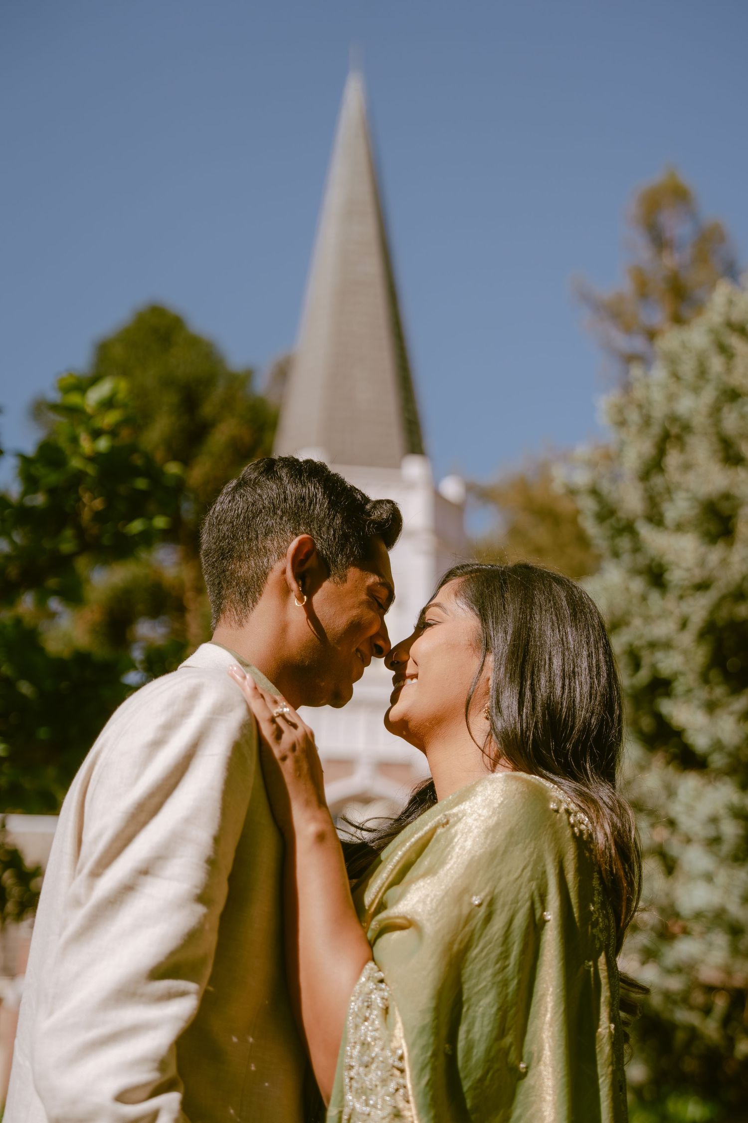 Couple in traditional South Asian attire sharing a romantic moment outdoors with a church steeple and trees behind them.