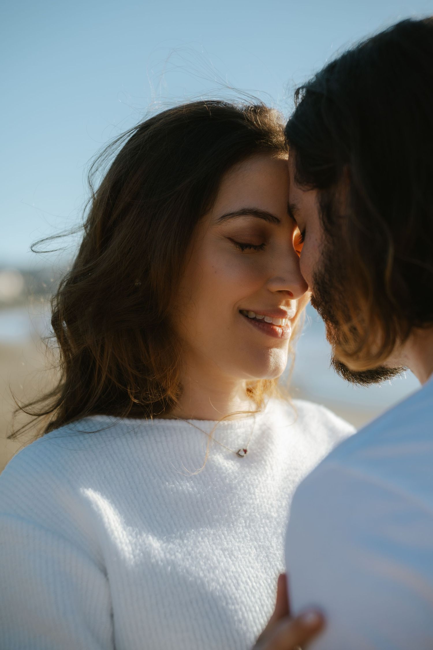 A couple shares a tender forehead-to-forehead moment outdoors, with the woman smiling softly in a white sweater.