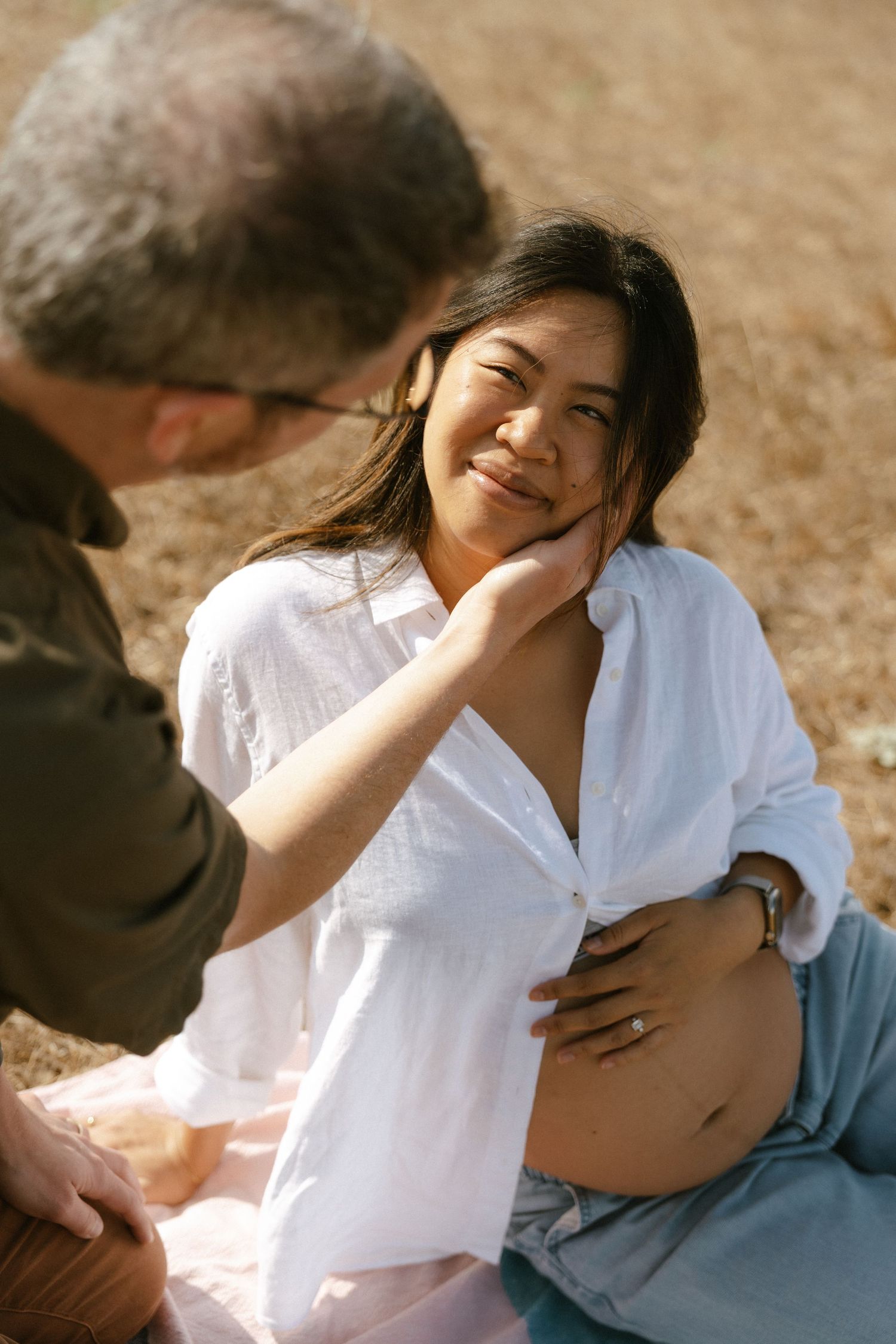 A man tenderly touches a pregnant woman's face as she smiles warmly while sitting outdoors in soft sunlight.
