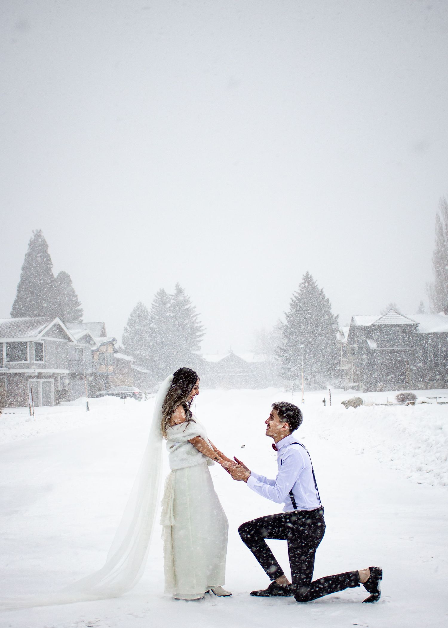 Groom kneeling in snow proposing to bride in white dress during winter snowstorm with snowy trees and houses backdrop.