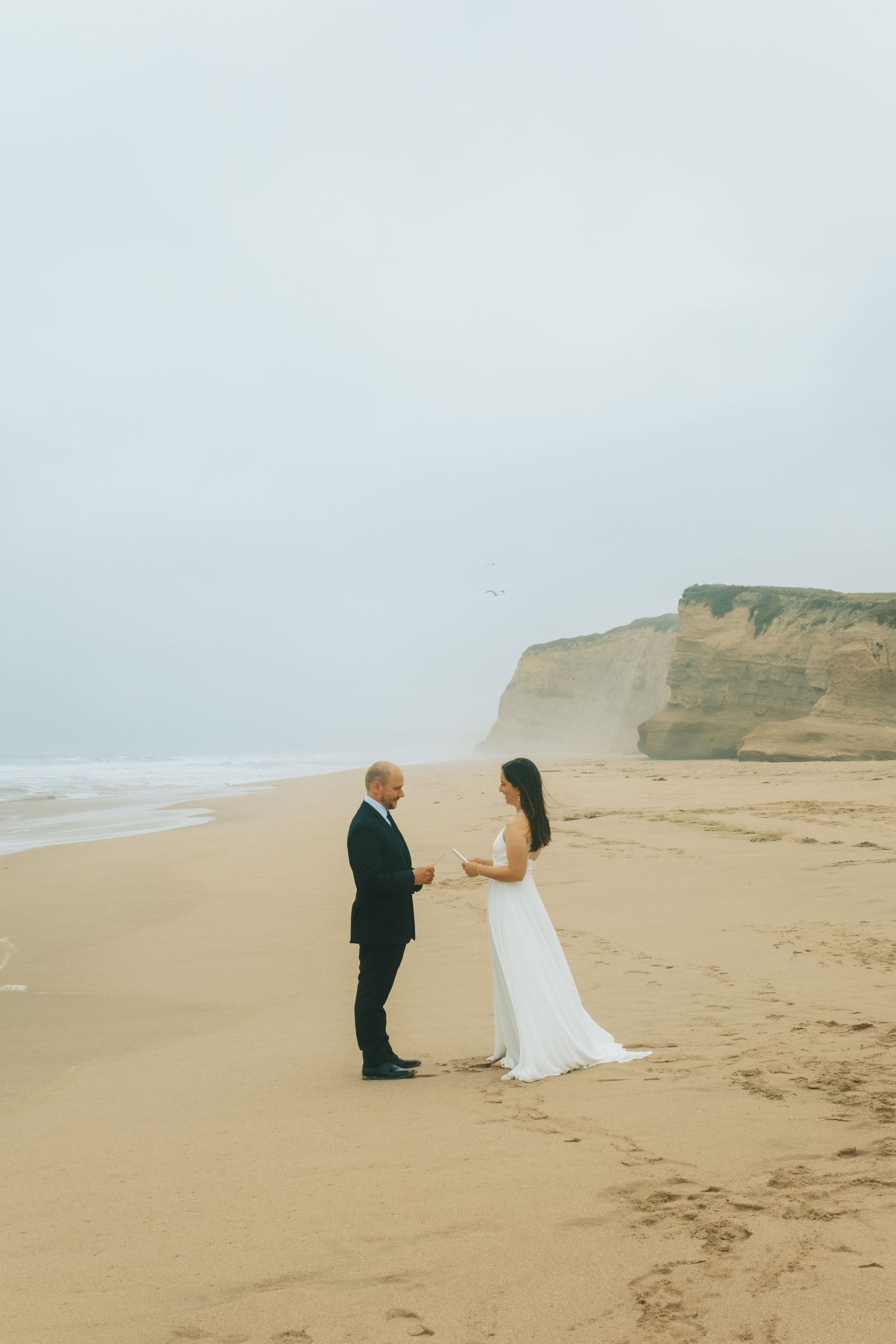 Couple exchanging vows on a misty beach, bride in white gown and groom in dark suit, with sandy shore and cliffs.
