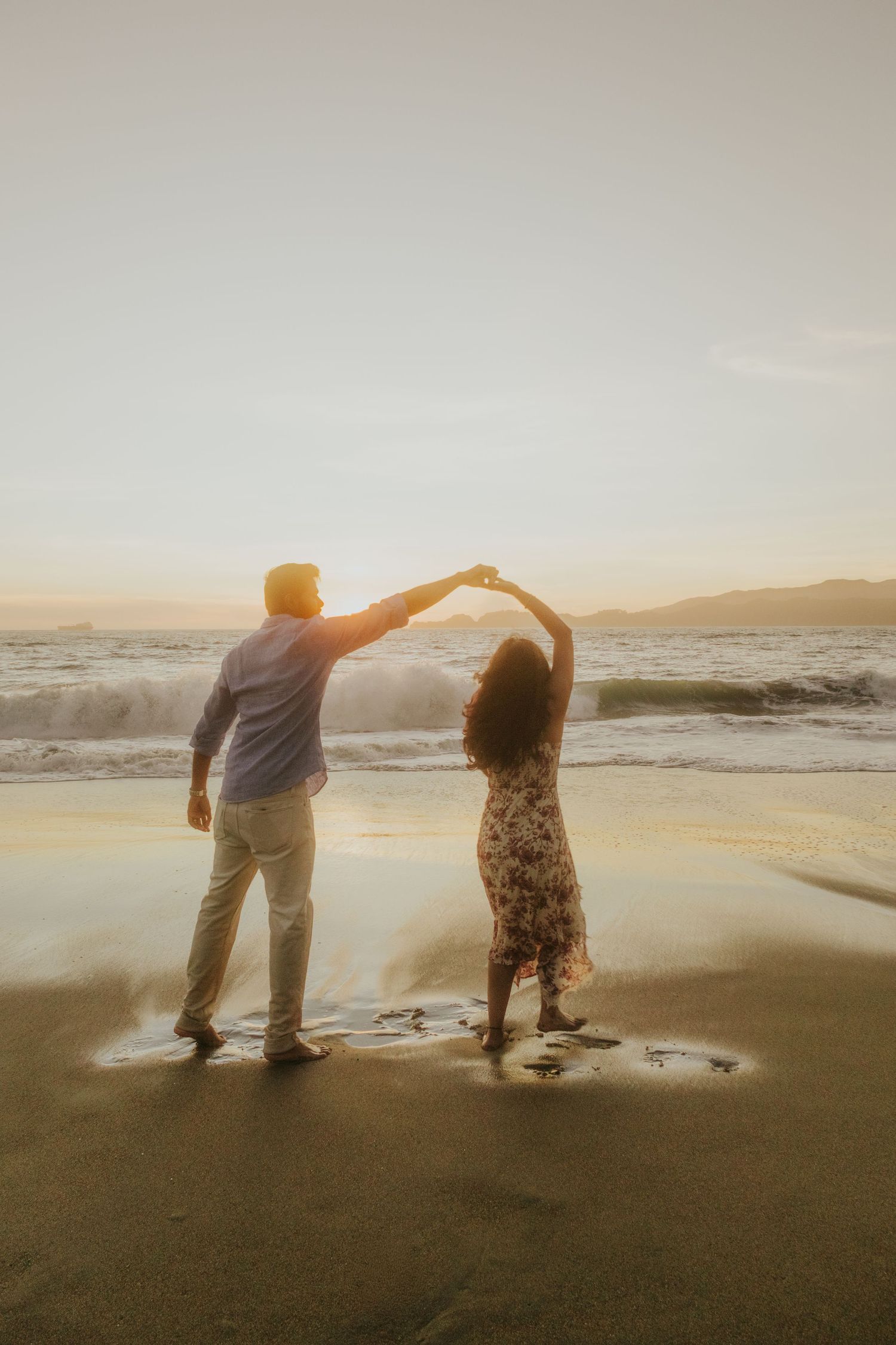 A couple dancing together on the beach at sunset with arms raised.
