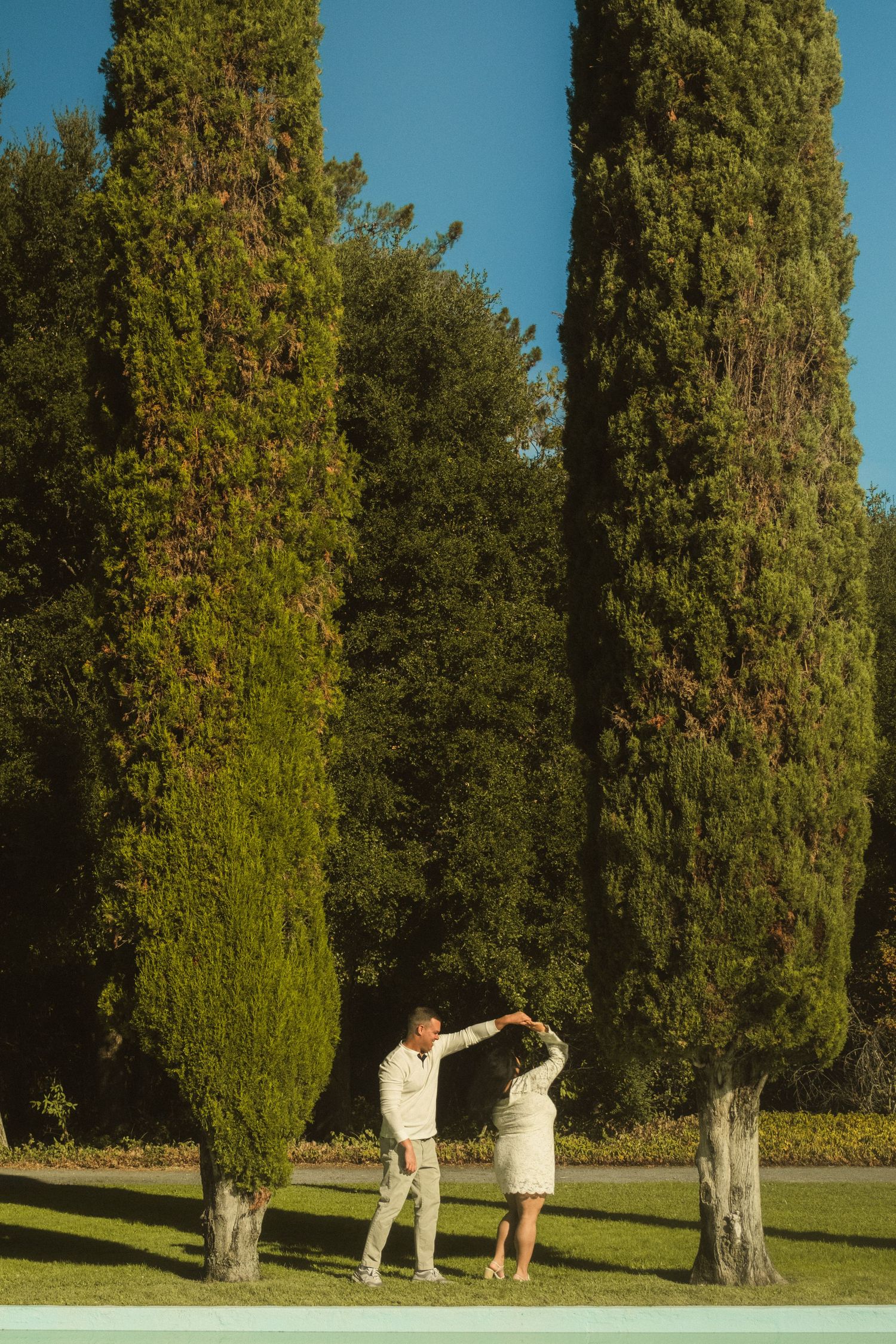 A couple dances playfully on a grassy lawn surrounded by cypress trees.