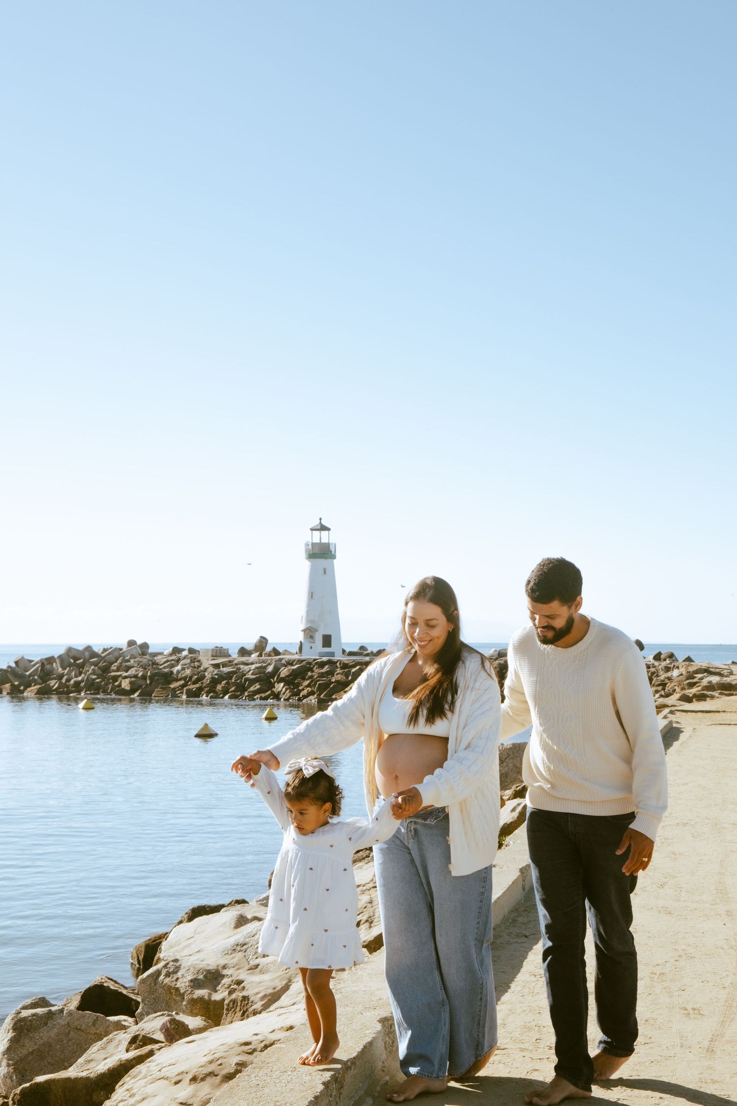 Family of three walking along rocky shoreline near a white lighthouse on a clear sunny day.
