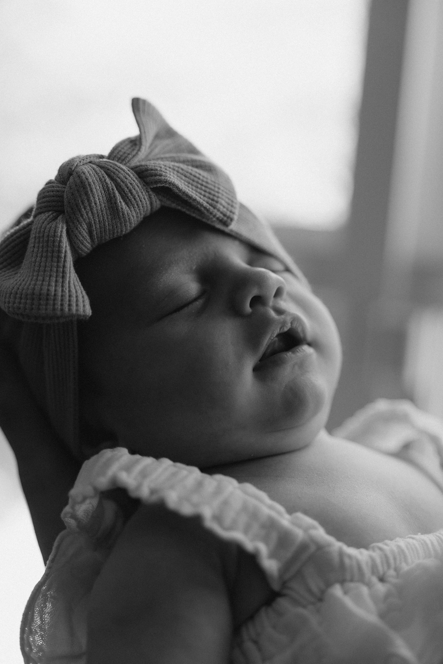 Black and white photo of a sleeping newborn baby wearing a bow headband, peacefully resting in someone's arms.