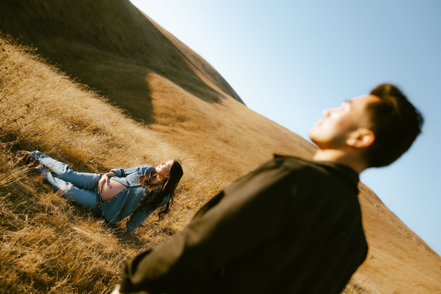 A woman relaxes on a golden hillside while a man in a dark jacket stands in the foreground under a clear blue sky.