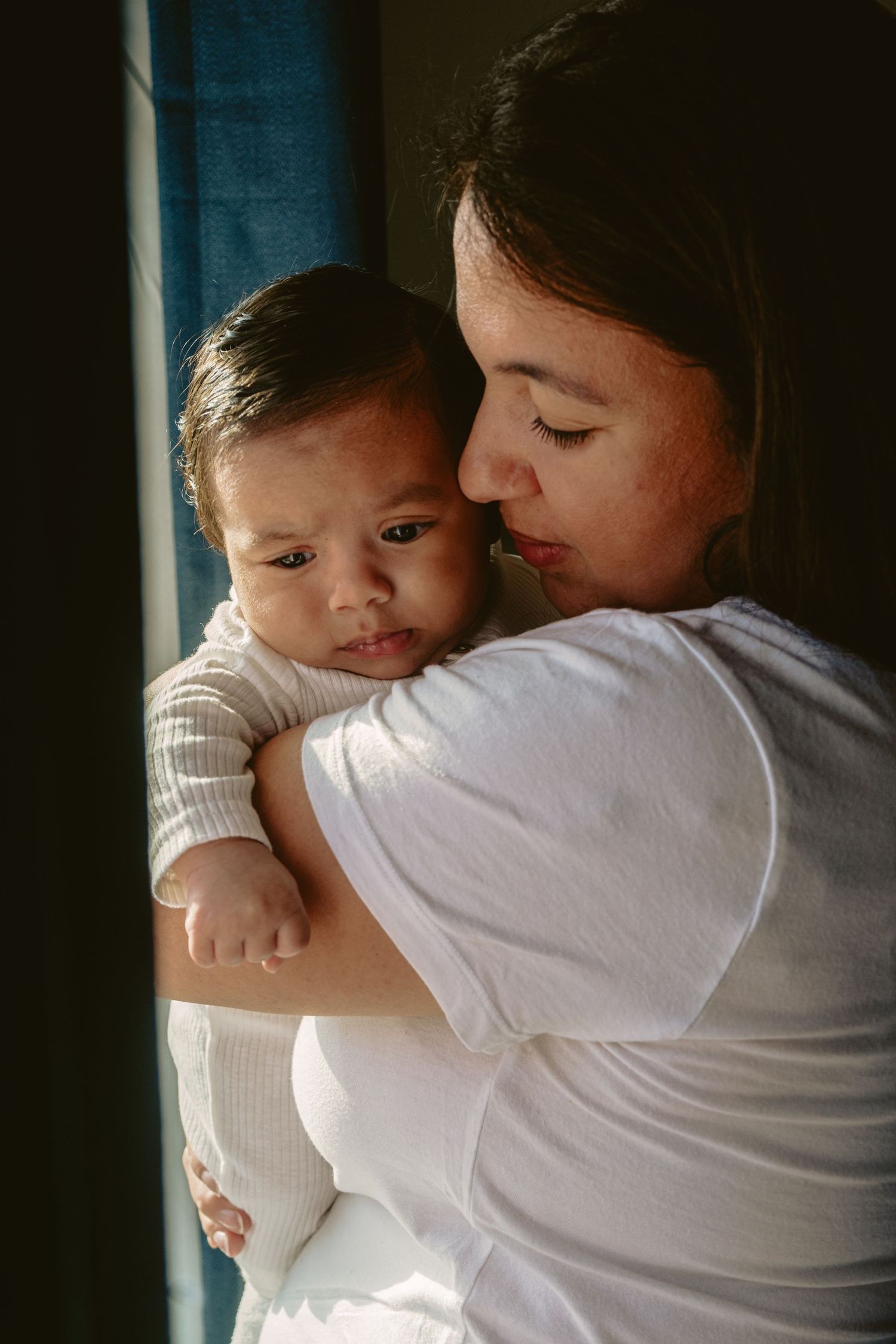 Mother in white shirt tenderly holding and kissing her newborn baby by a sunlit window.