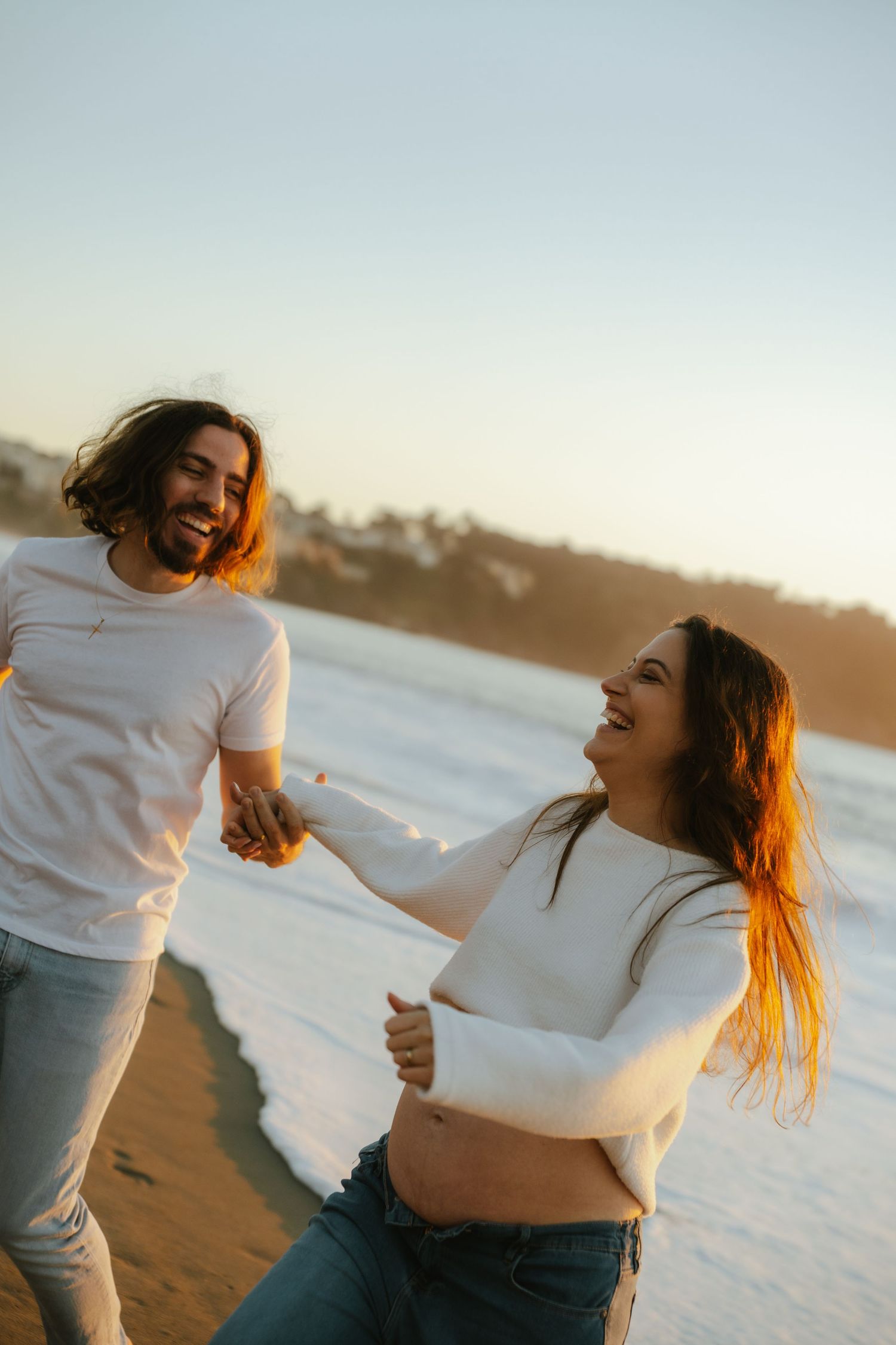 Happy couple holding hands and laughing on a sunny beach at golden hour, wearing matching white tops and jeans.