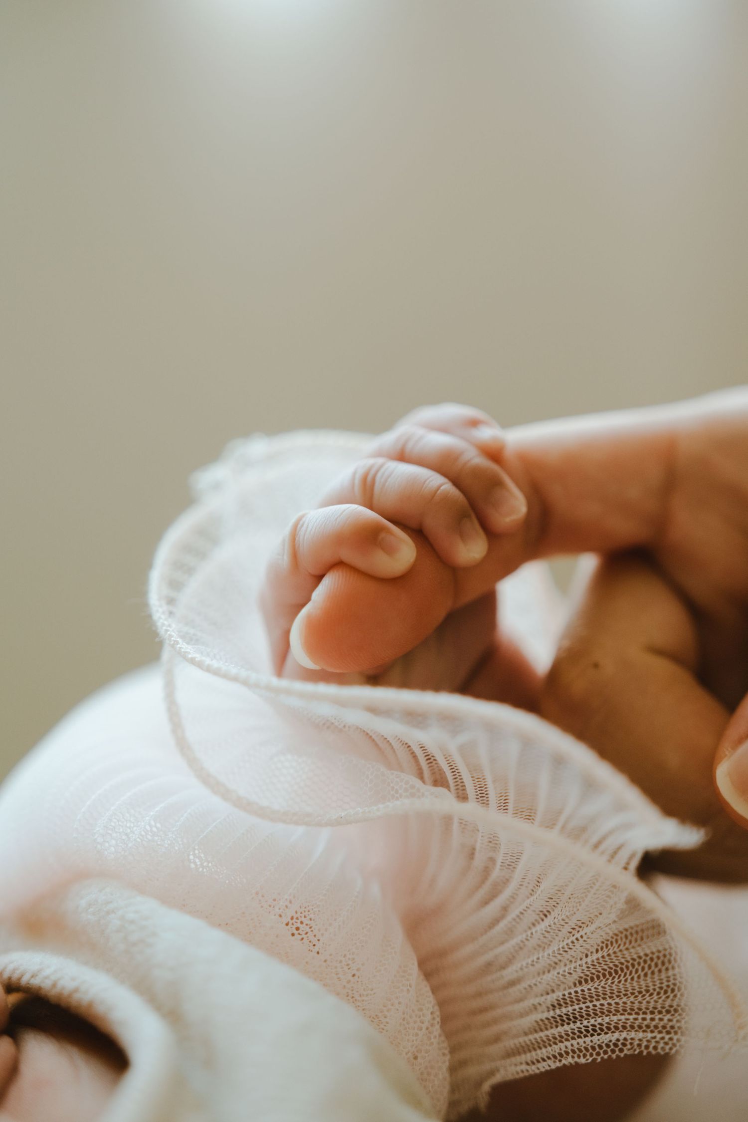 A newborn baby's tiny hand gently gripping an adult's finger, dressed in a white lace outfit.