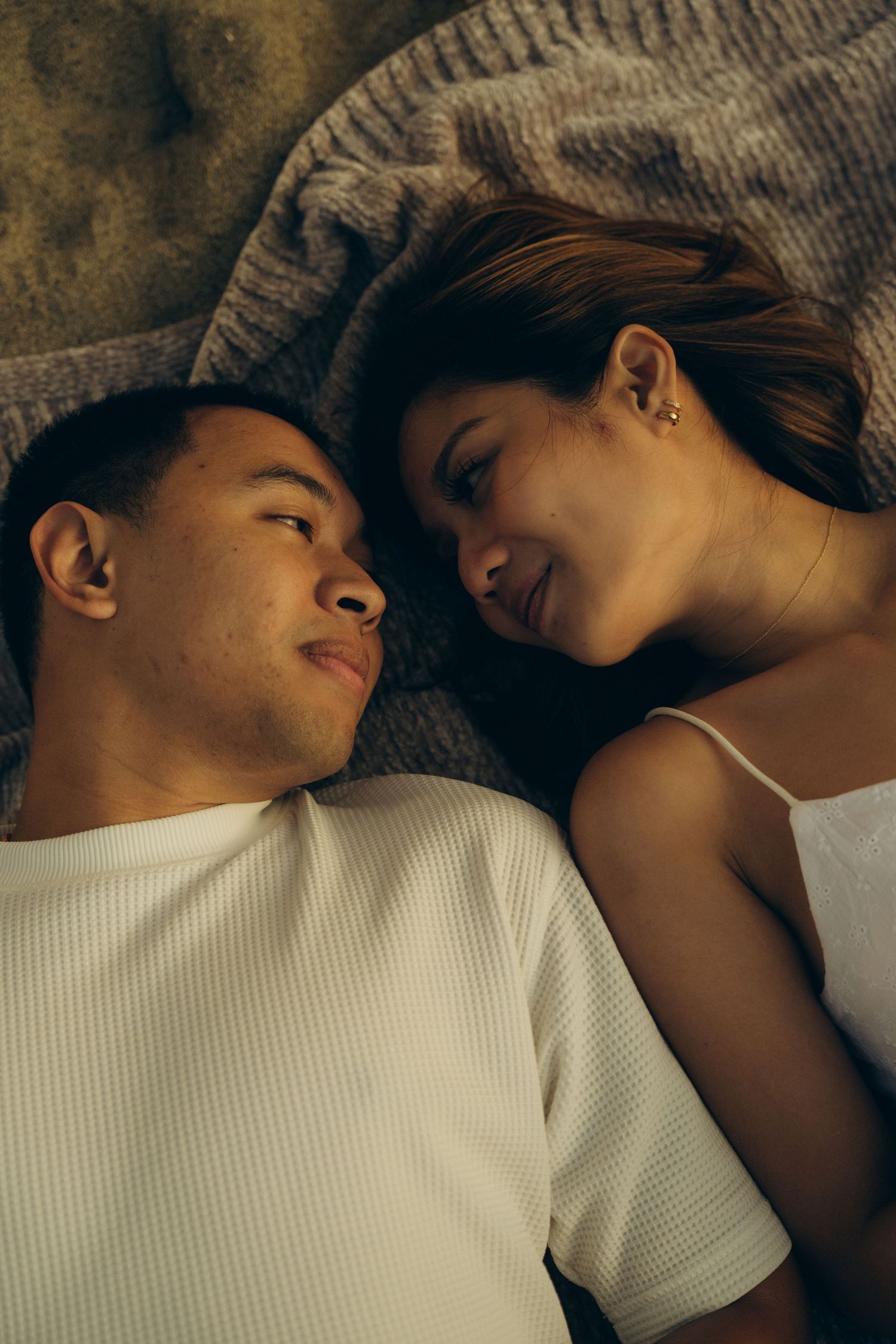 A young couple lying on a bed, gazing at each other with warm smiles in a cozy, intimate setting.