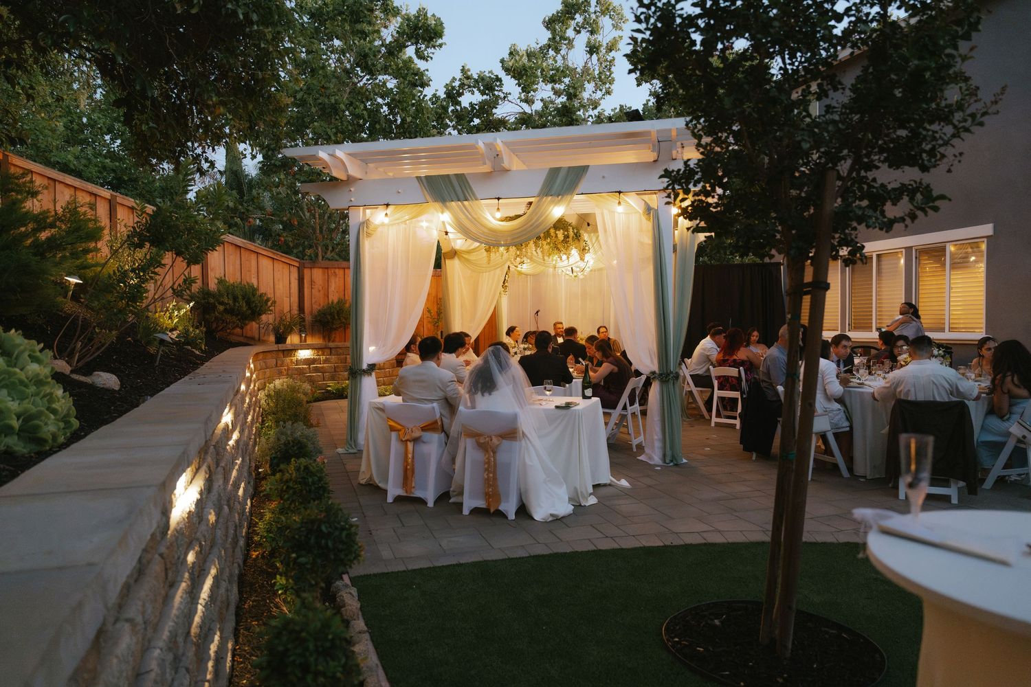 Elegant outdoor backyard wedding reception under a white pergola with draped fabric and floral decor at dusk.
