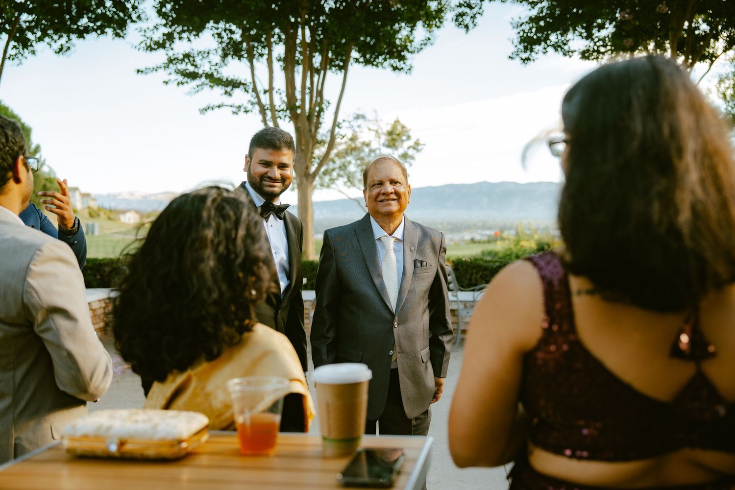 Two men in formal attire smile and greet guests at an outdoor wedding reception with scenic hills in the background.