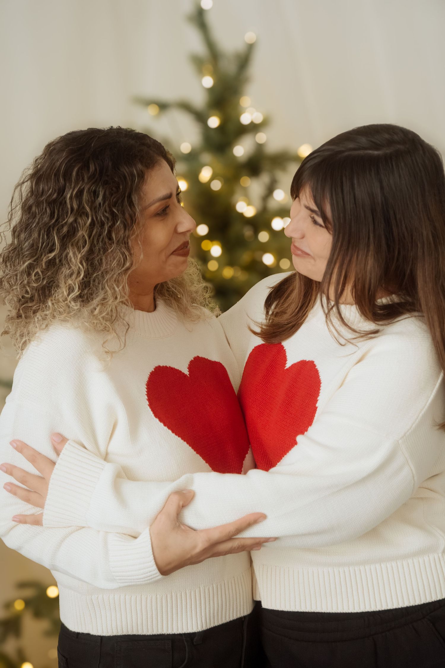 Friends embracing in matching white sweaters with red heart accents near a Christmas tree.