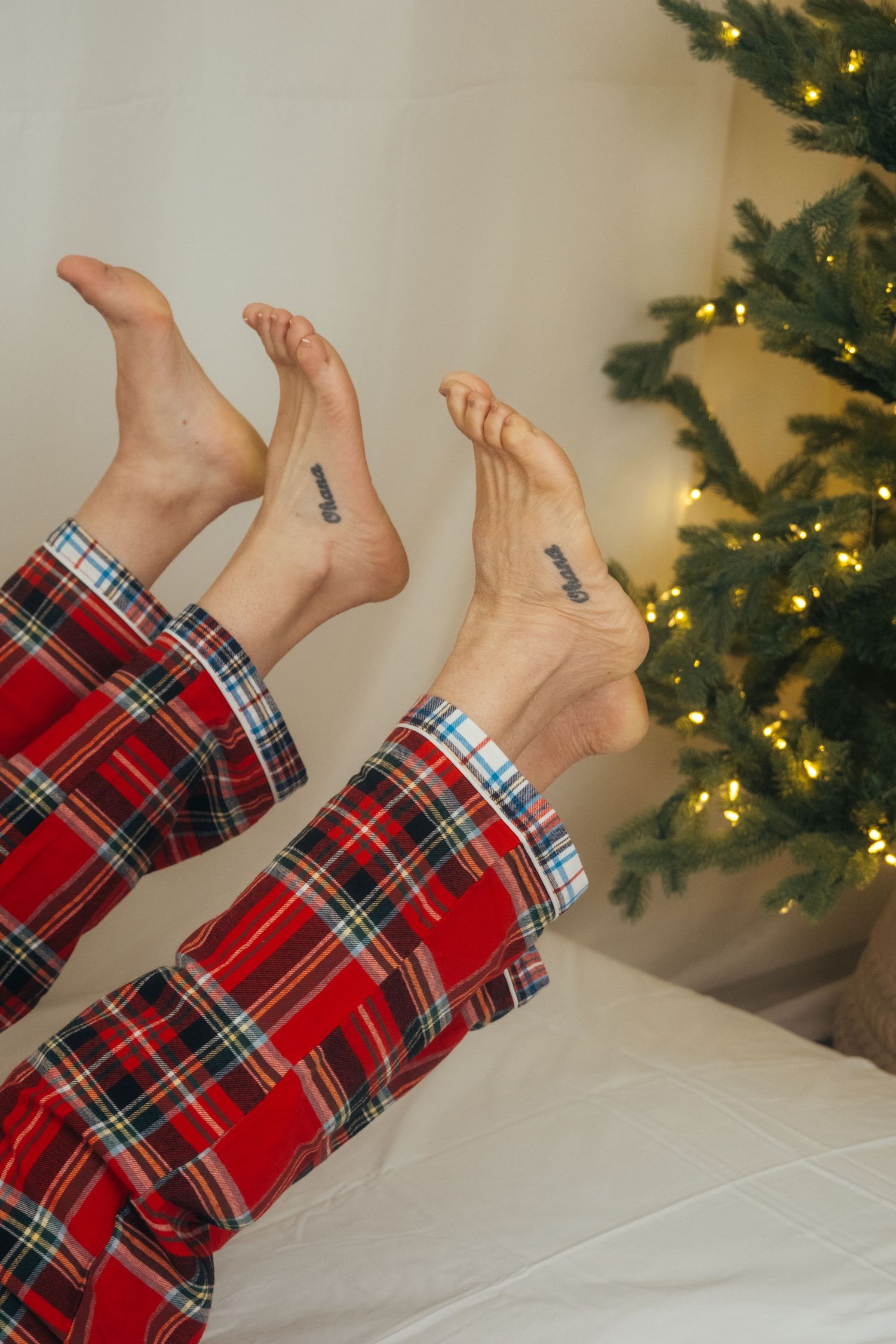 Bare feet with matching tattoos wearing plaid pajama bottoms against Christmas tree background.